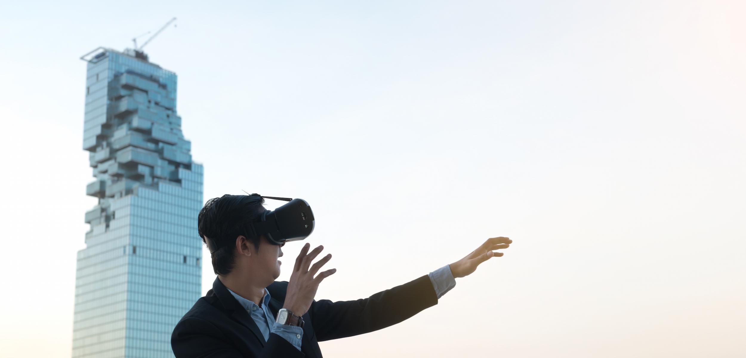 A man in a suit is wearing a virtual reality headset and reaching out with one hand, standing against a modern skyscraper. The sky is light and clear.