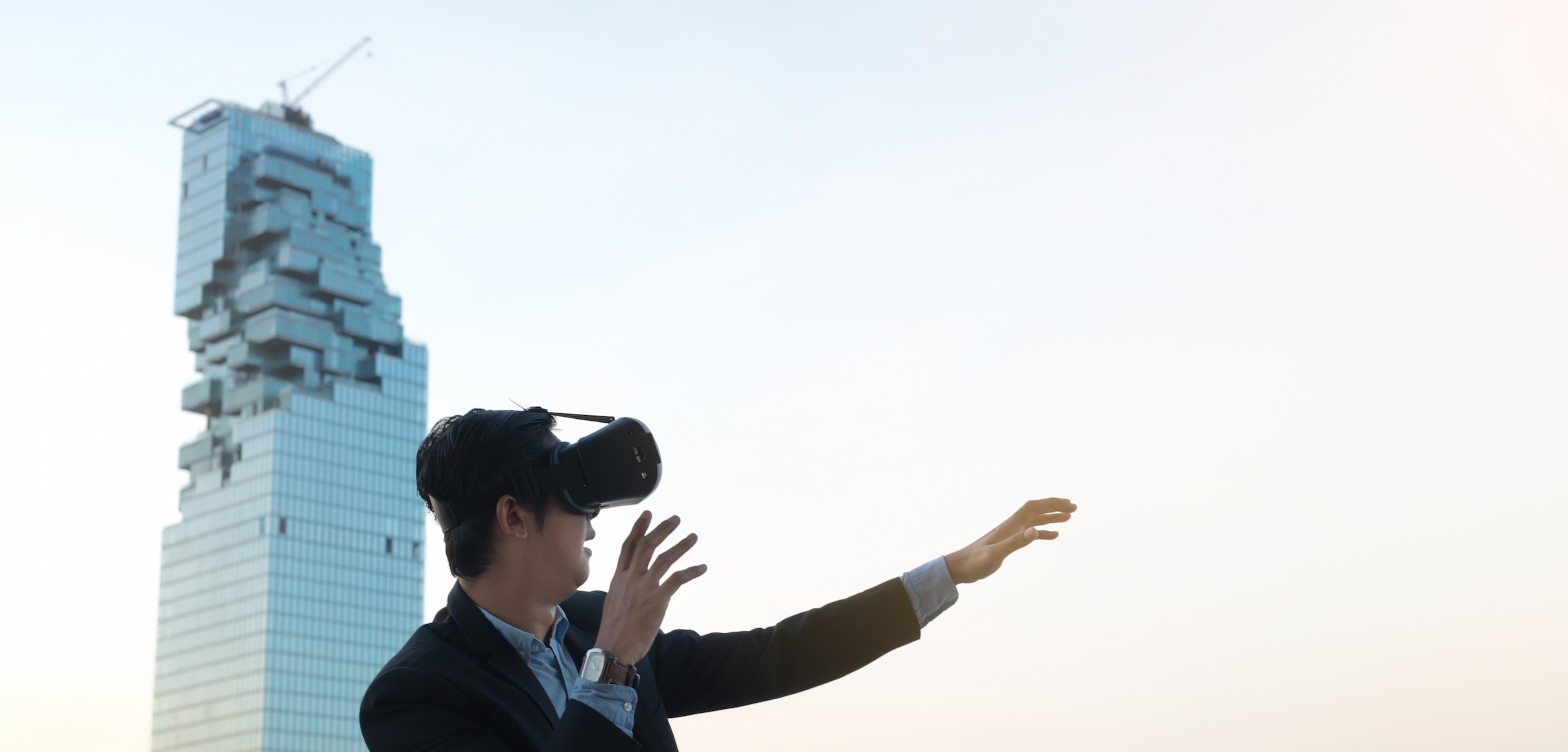 A man in a suit is wearing a virtual reality headset and reaching out with one hand, standing against a modern skyscraper. The sky is light and clear.