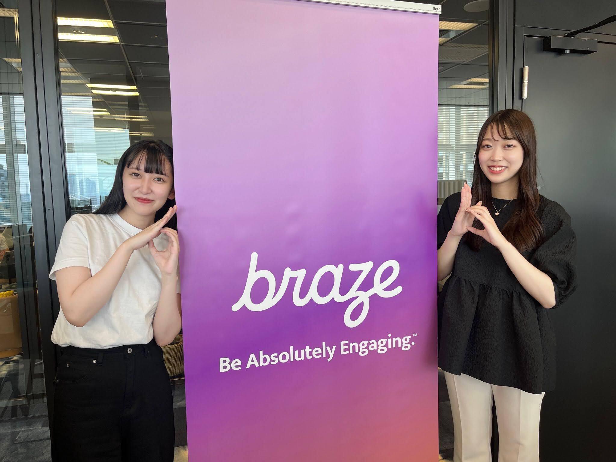 ブライズのバナーの前でポーズをとる二人の女性。バナーには「Be Absolutely Engaging」と書かれている。