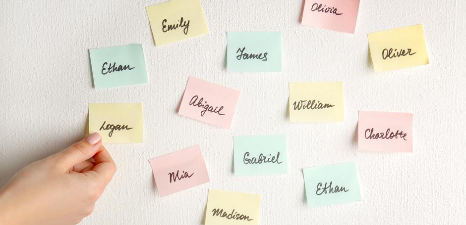 A hand places a sticky note on a wall filled with colorful notes featuring names written in cursive. The notes are in pastel colors, including pink, blue, and yellow.