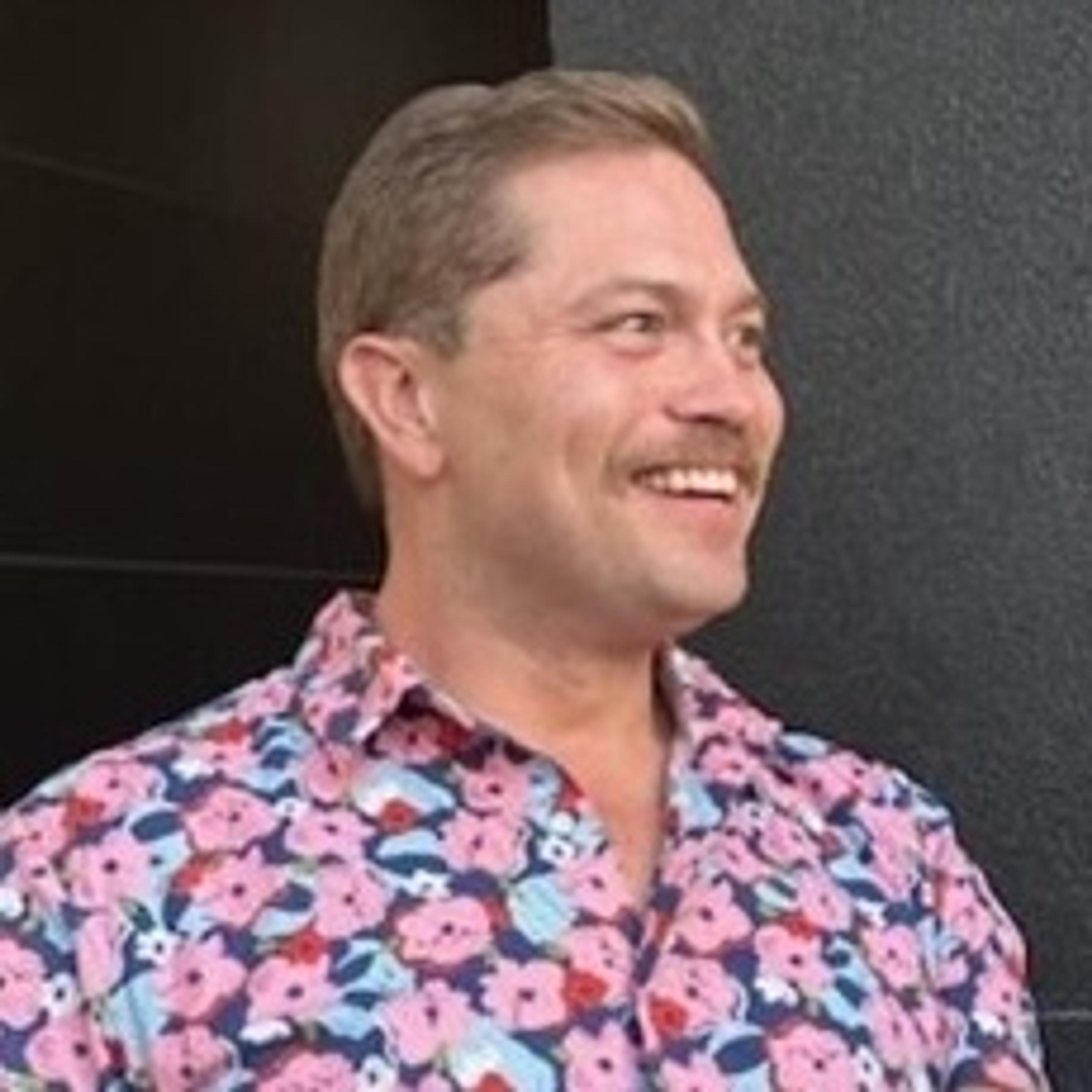 Smiling person with short light-brown hair and a mustache wearing a colorful pink floral shirt, looking to the side against a dark background.