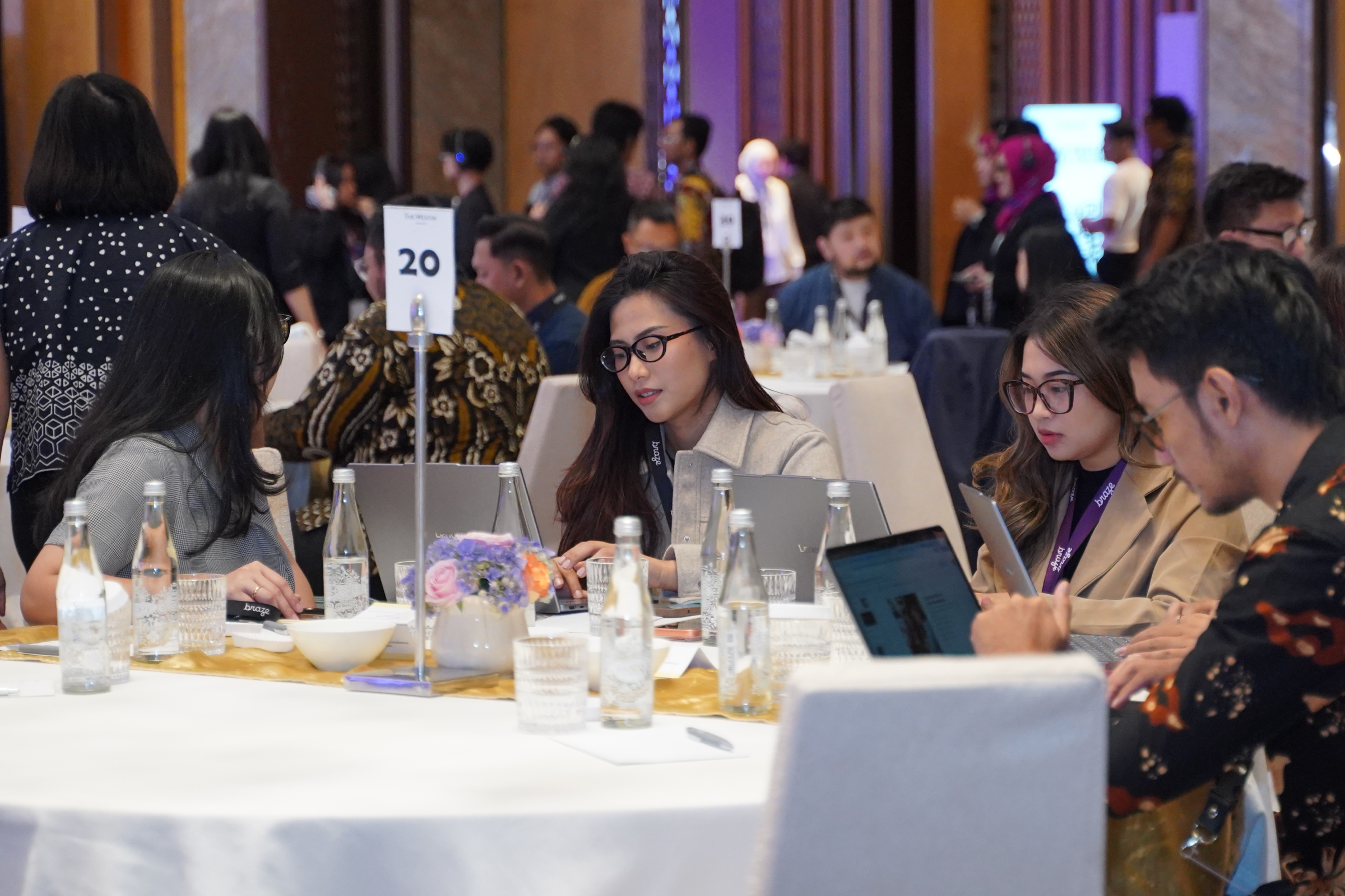 Conference attendees work on laptops and tablets around a banquet table marked with the number 20.