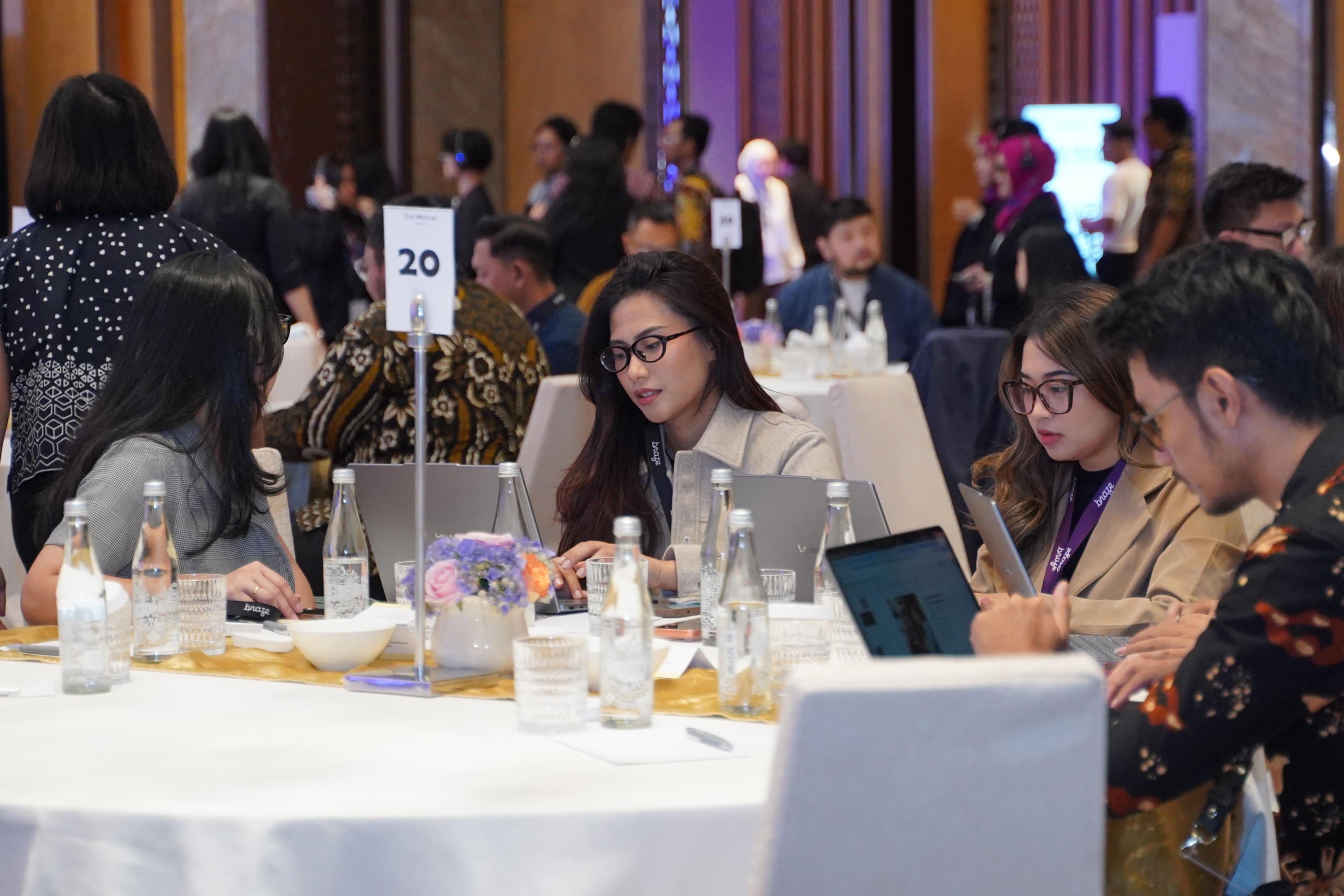 Conference attendees work on laptops and tablets around a banquet table marked with the number 20.