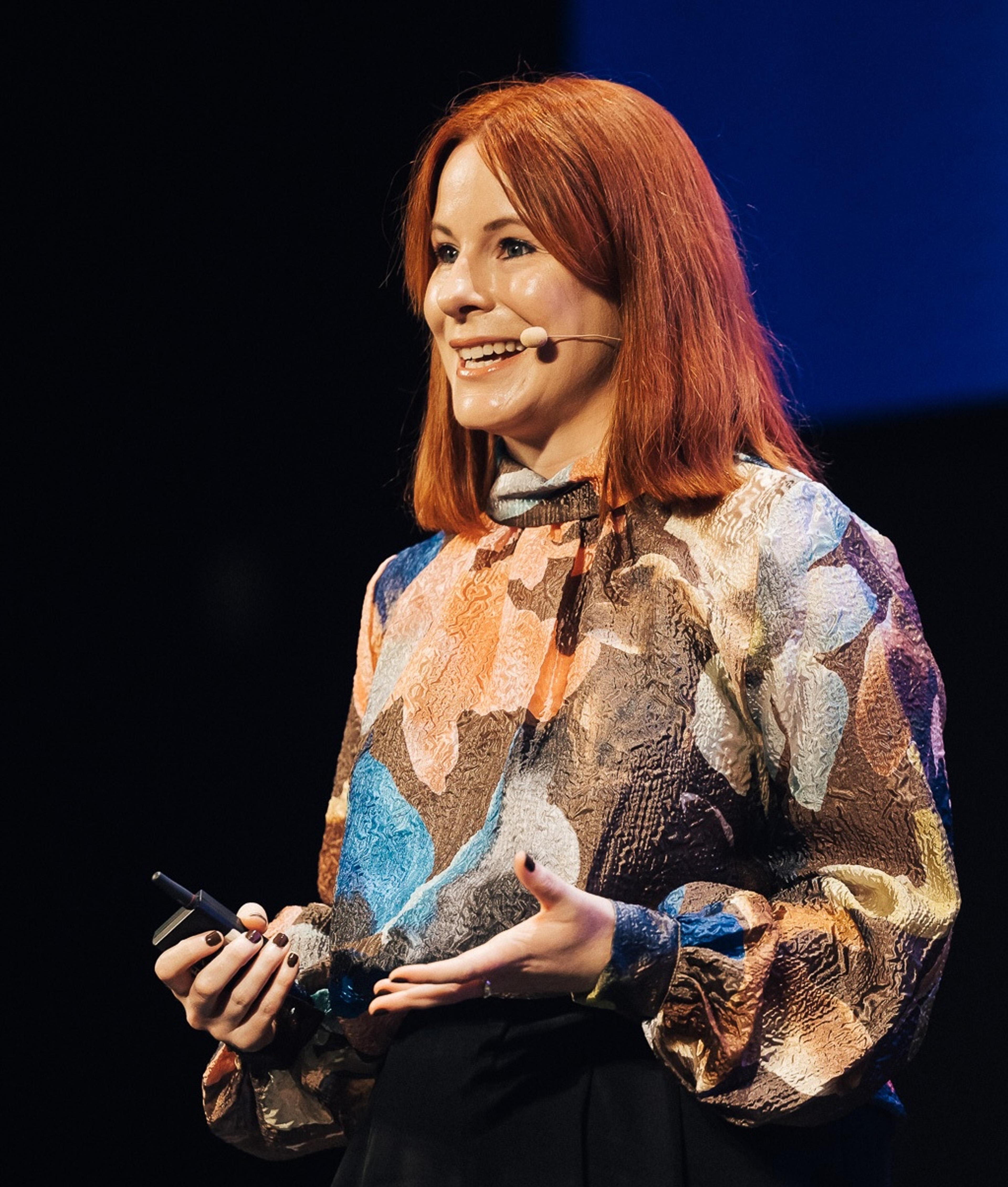 Smiling red-haired woman wearing a headset microphone and colorful patterned blouse speaks onstage while holding a presentation remote.