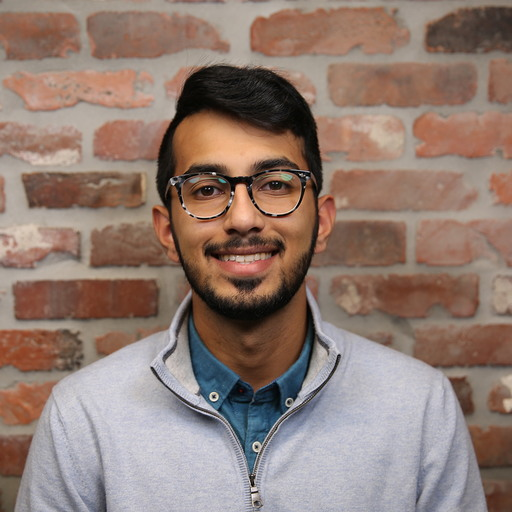 A young man with glasses smiles in front of a brick wall. He is wearing a light gray sweater over a blue shirt.