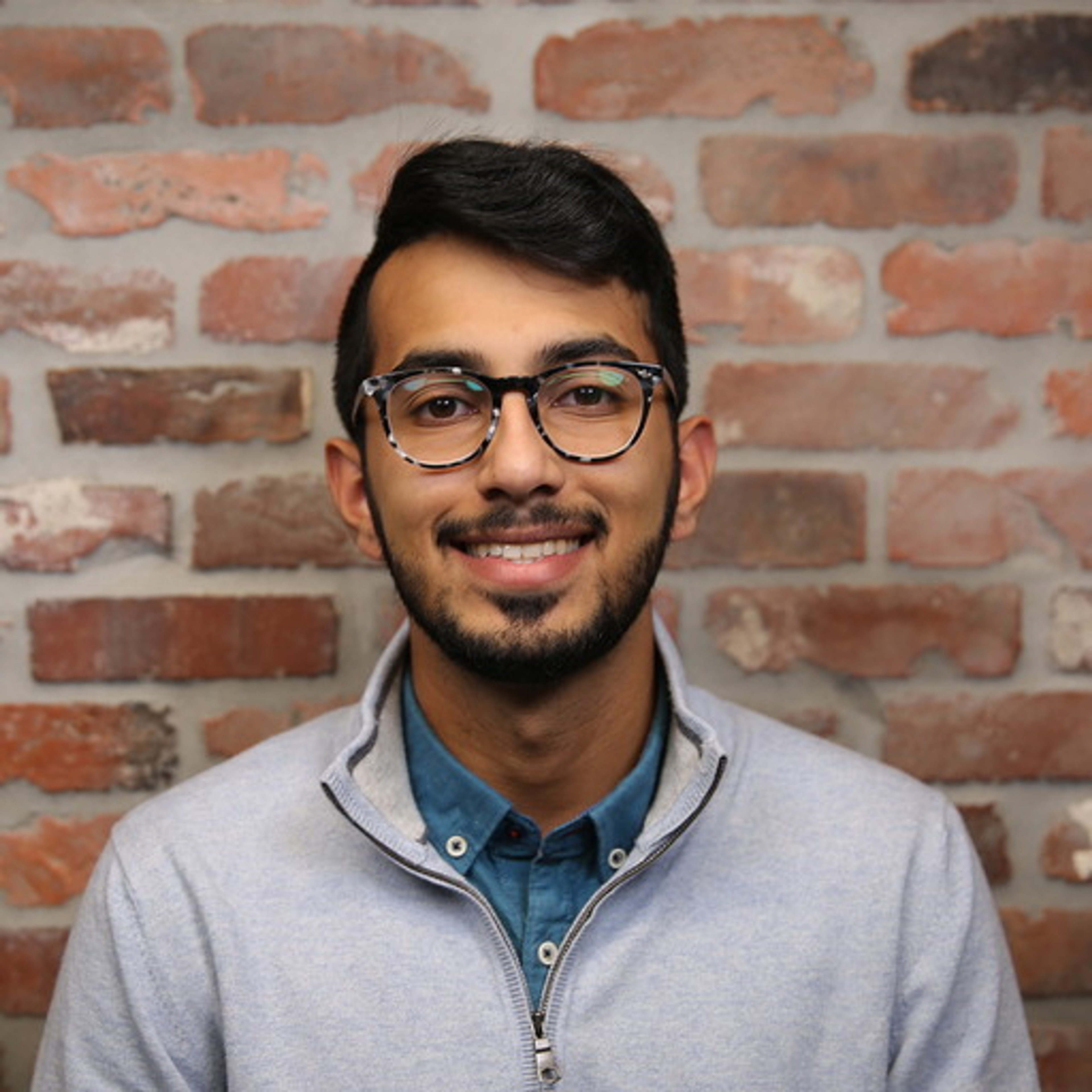 A young man with glasses smiles in front of a brick wall. He is wearing a light gray sweater over a blue shirt.