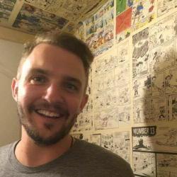 A smiling man with short hair stands in front of a wall covered in comic strip art.