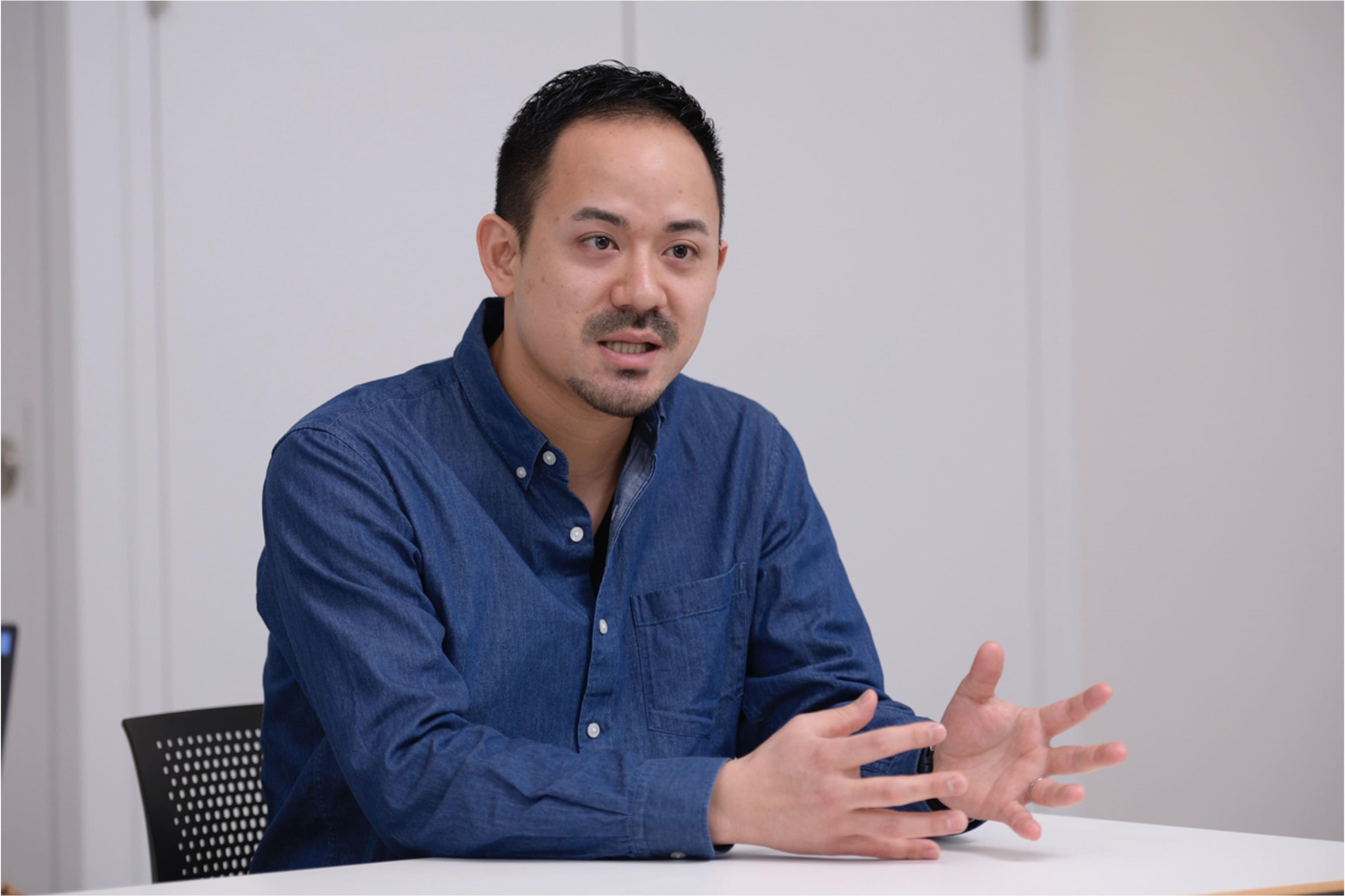男性がテーブルに座り、話している様子。青いシャツを着ており、手を使って説明しています。