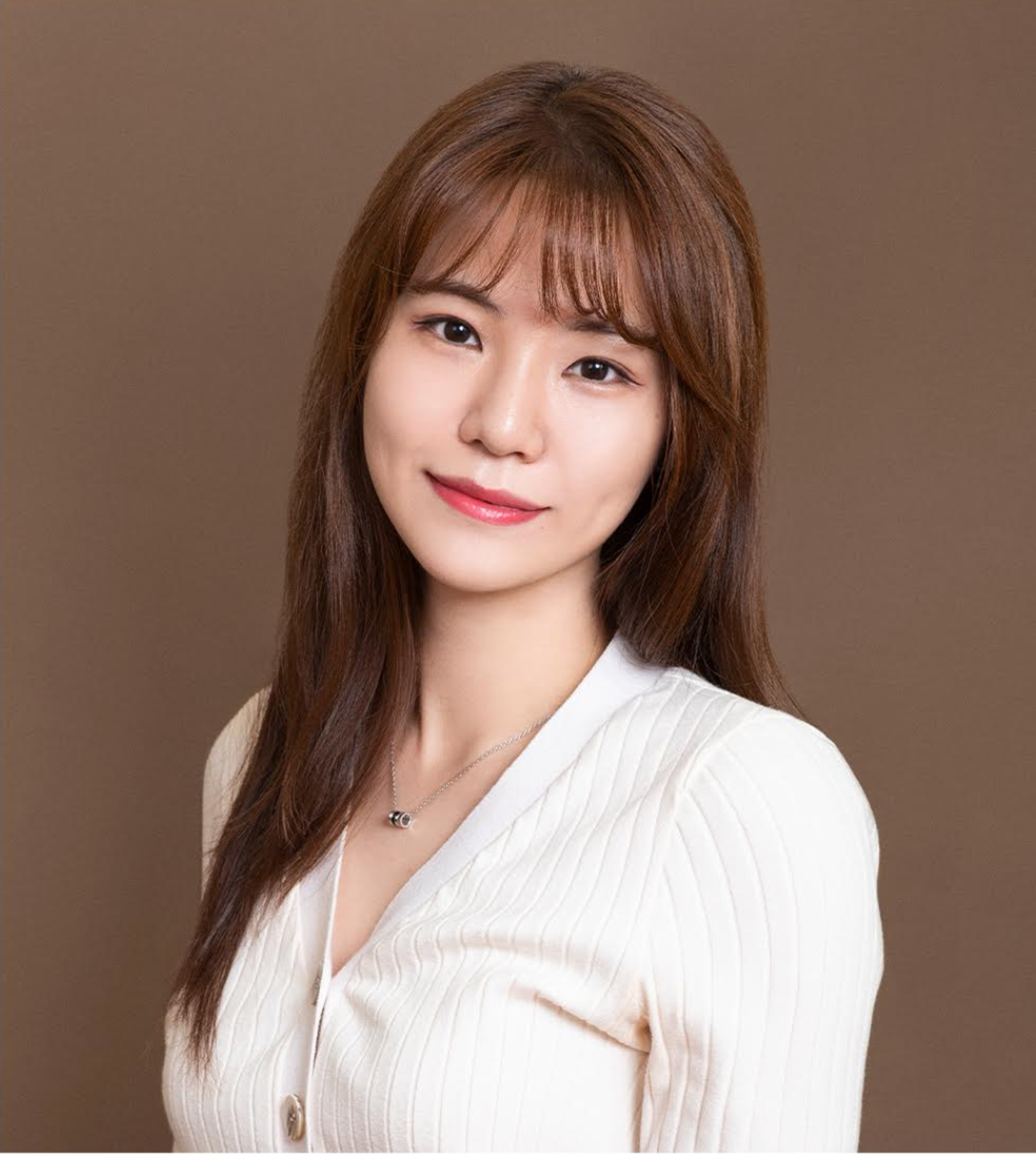 A woman with long brown hair smiles softly, wearing a white ribbed sweater against a brown background. She has a delicate necklace and a natural makeup look.
