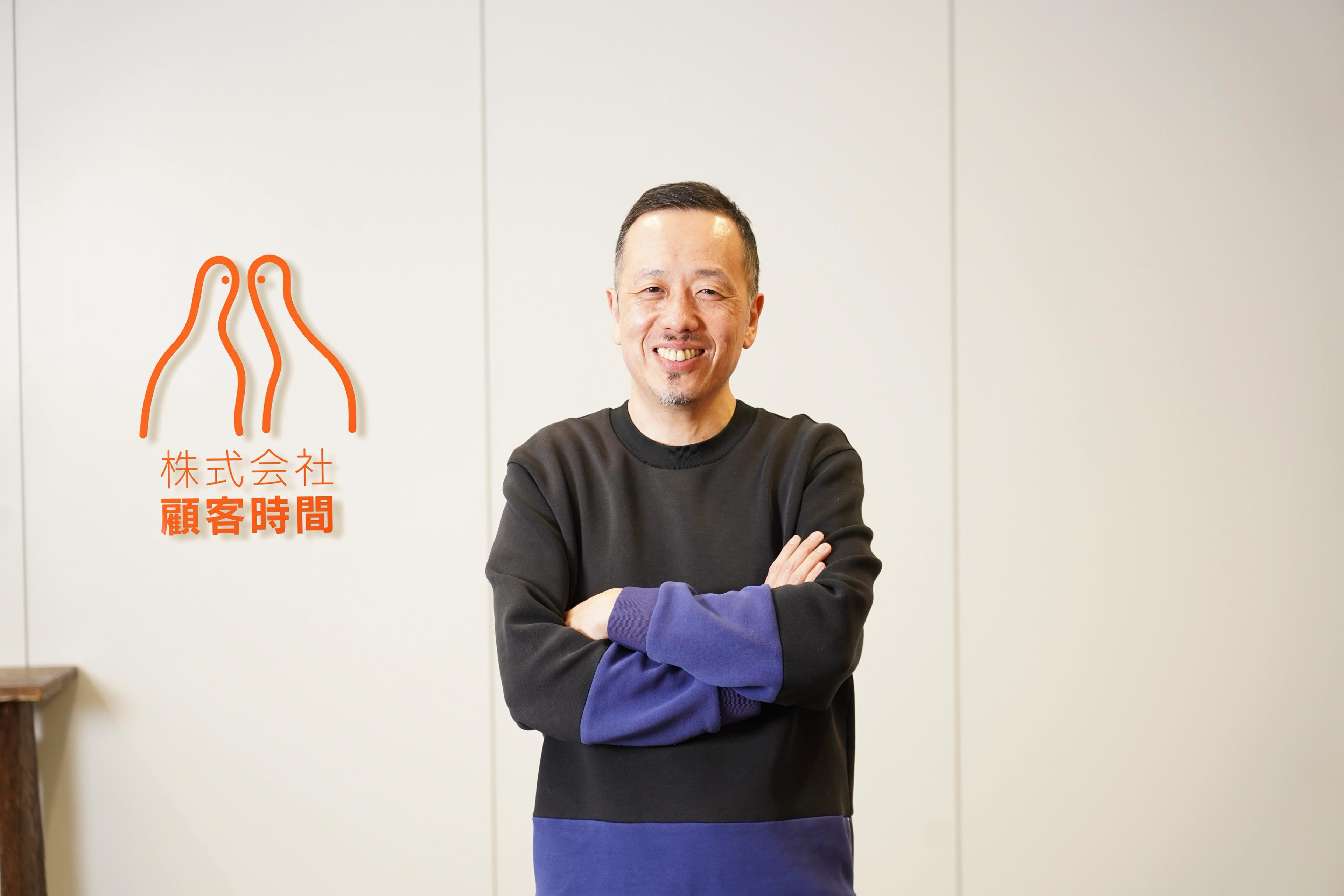腕を組んだ笑顔の男性が、白い壁に描かれたオレンジ色のロゴと日本語の文字の前に立っている。