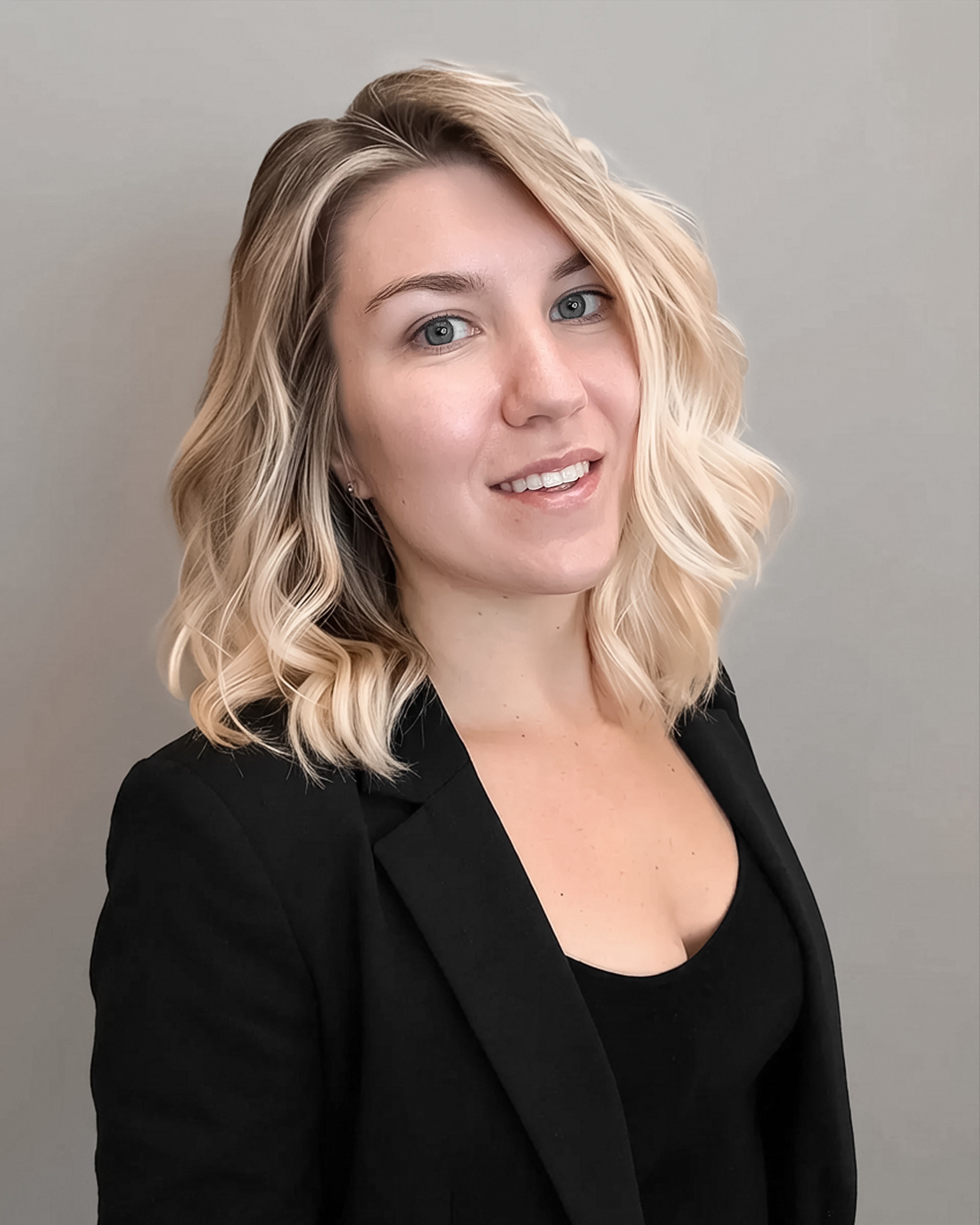 A woman with wavy blonde hair smiles at the camera, wearing a black blazer over a black top. The background is a light gray color.