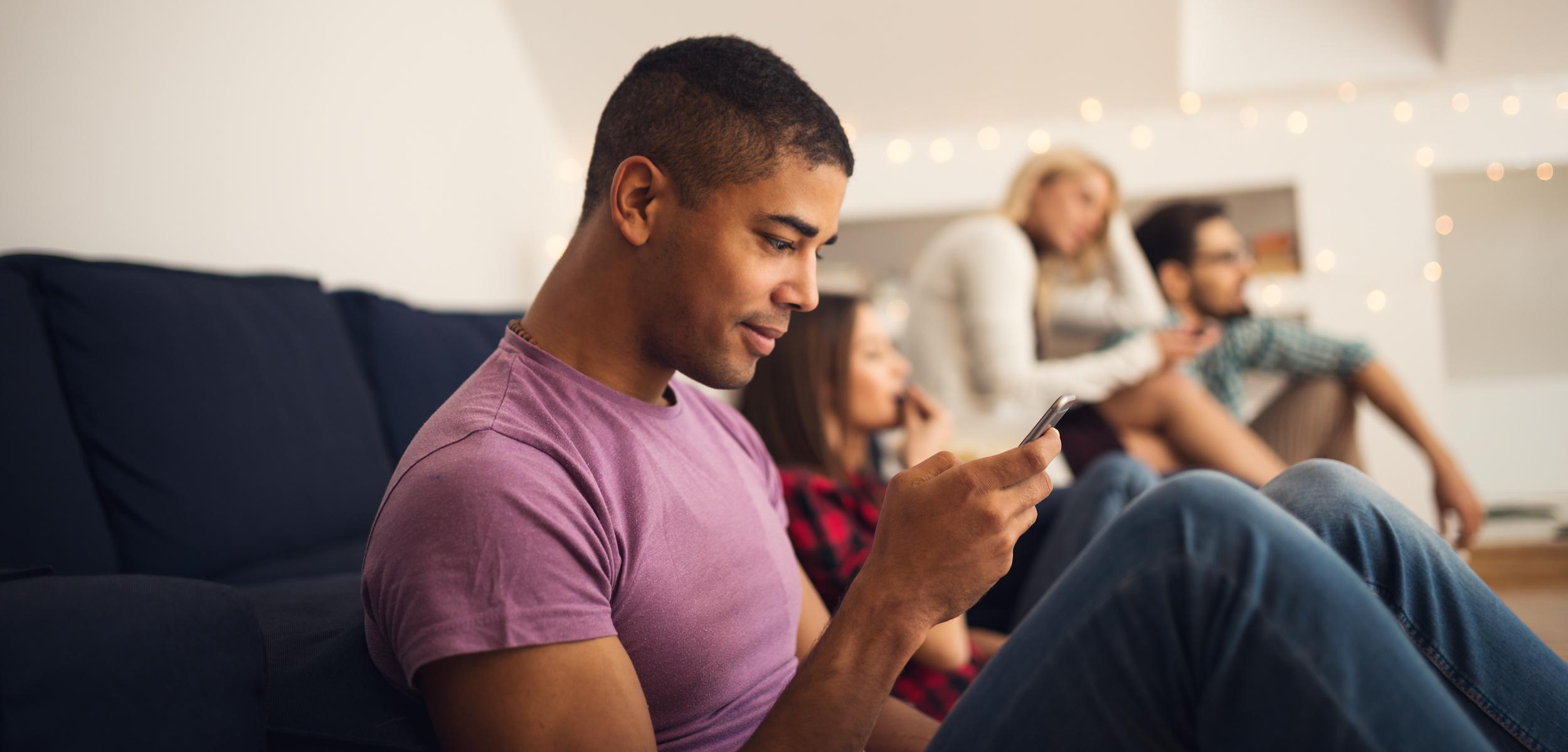 A young man sits on a couch, looking at his phone while two women and another man are seated in the background, engaged in conversation. The setting is cozy with soft lighting.