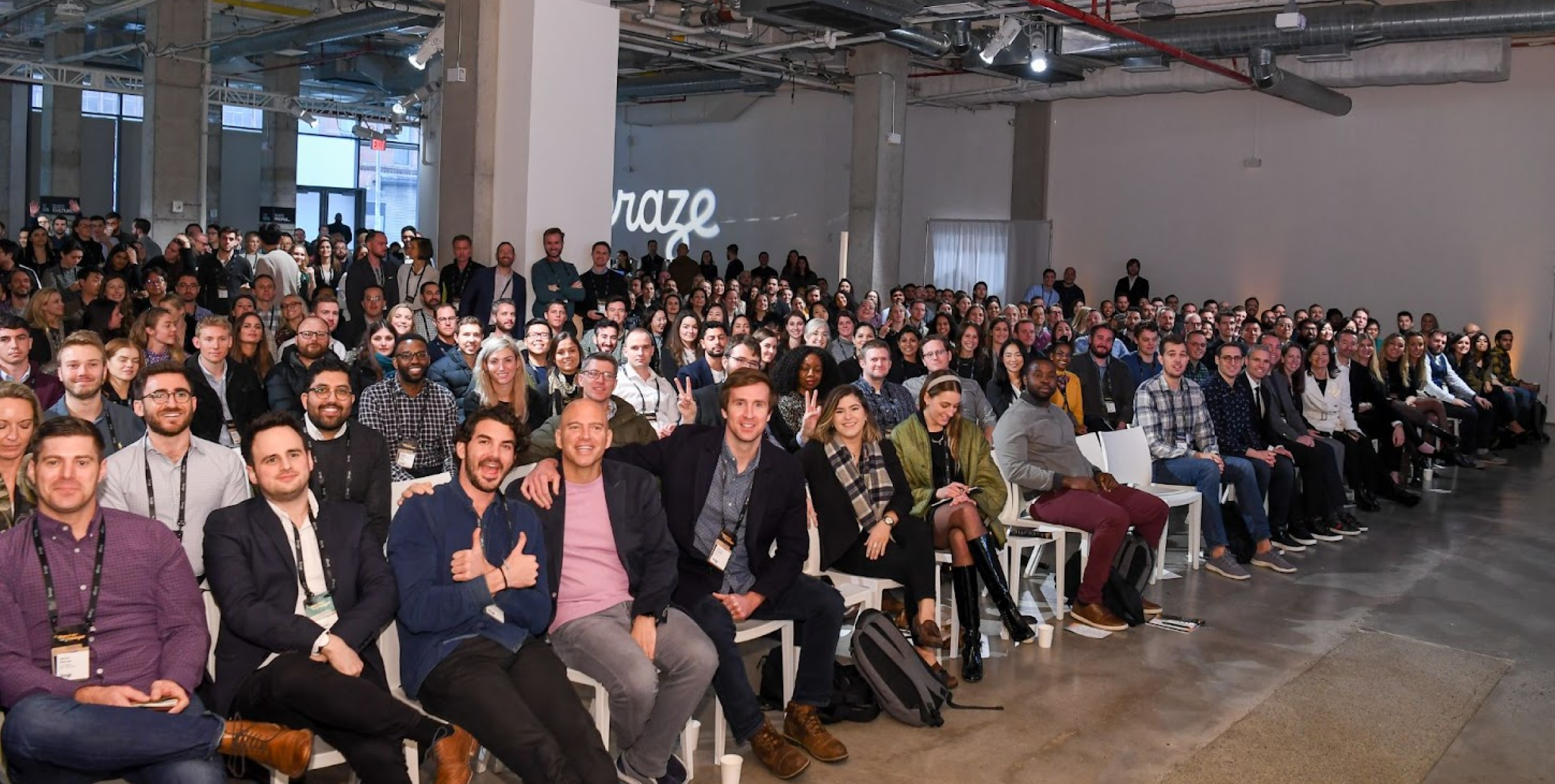 A large audience of diverse individuals seated in a spacious venue, facing the camera. The crowd appears engaged and attentive, with some people smiling and posing for the photo.