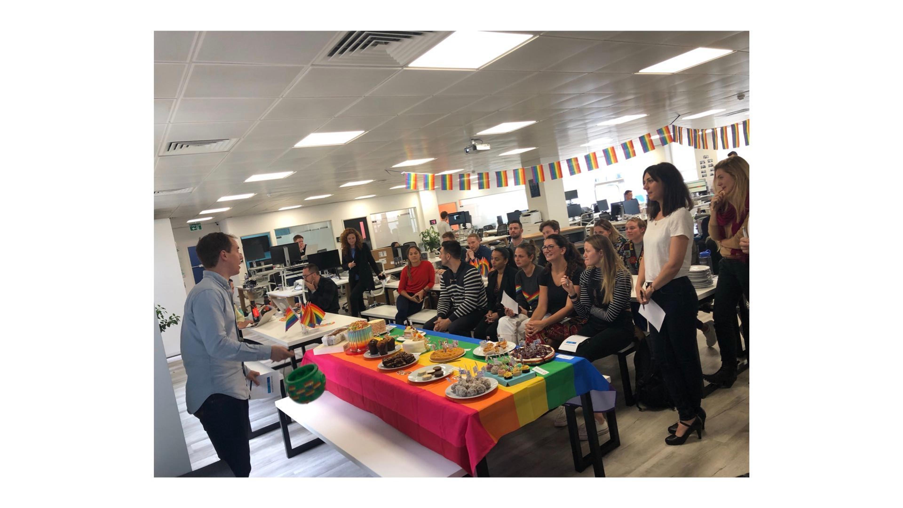 A diverse group of people gathers in an office space for a celebration, with a table decorated in rainbow colors and various desserts. Banners and flags are displayed in the background, creating a festive atmosphere.