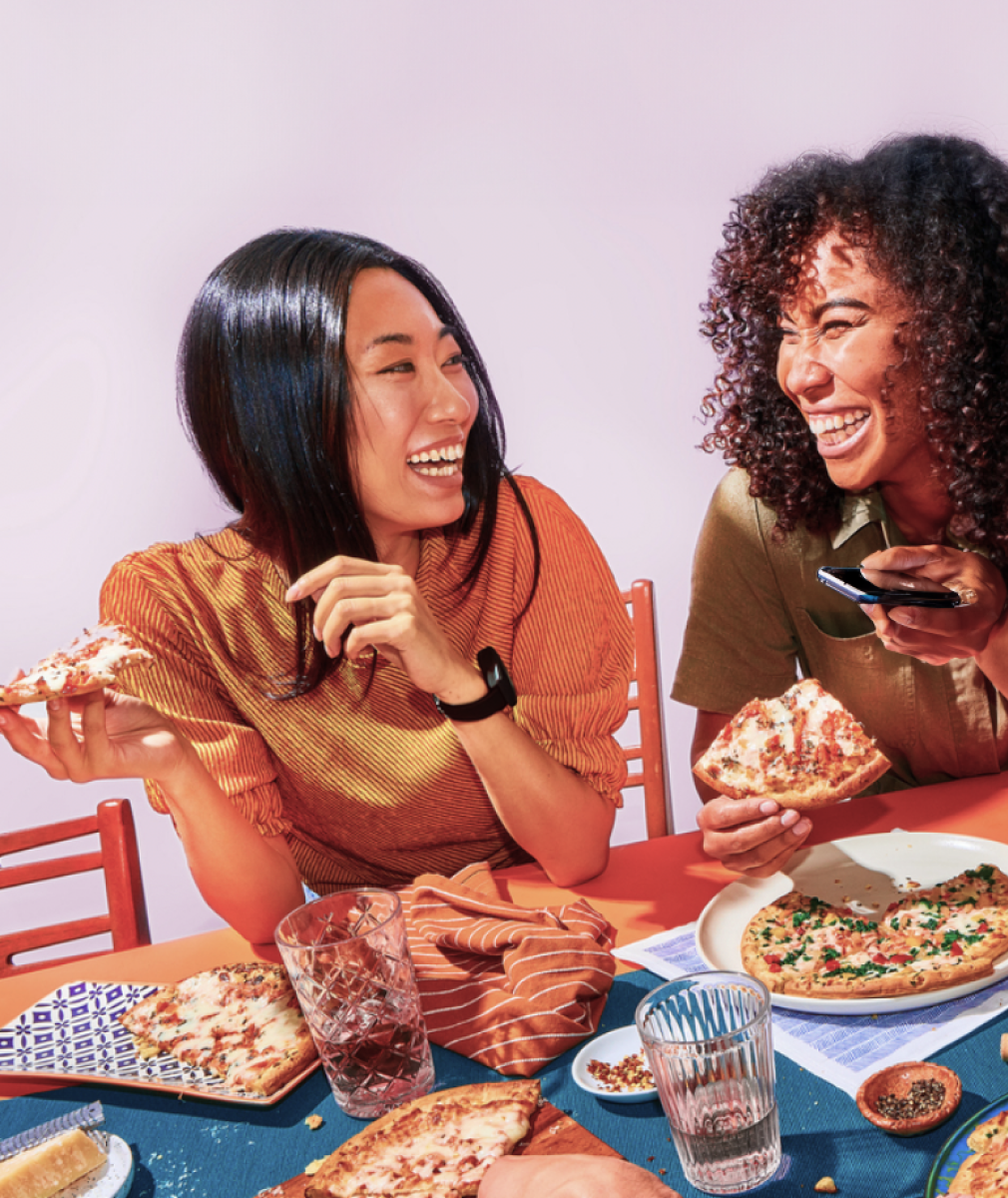 Two friends sit at a colorful table, laughing and eating pizza together.