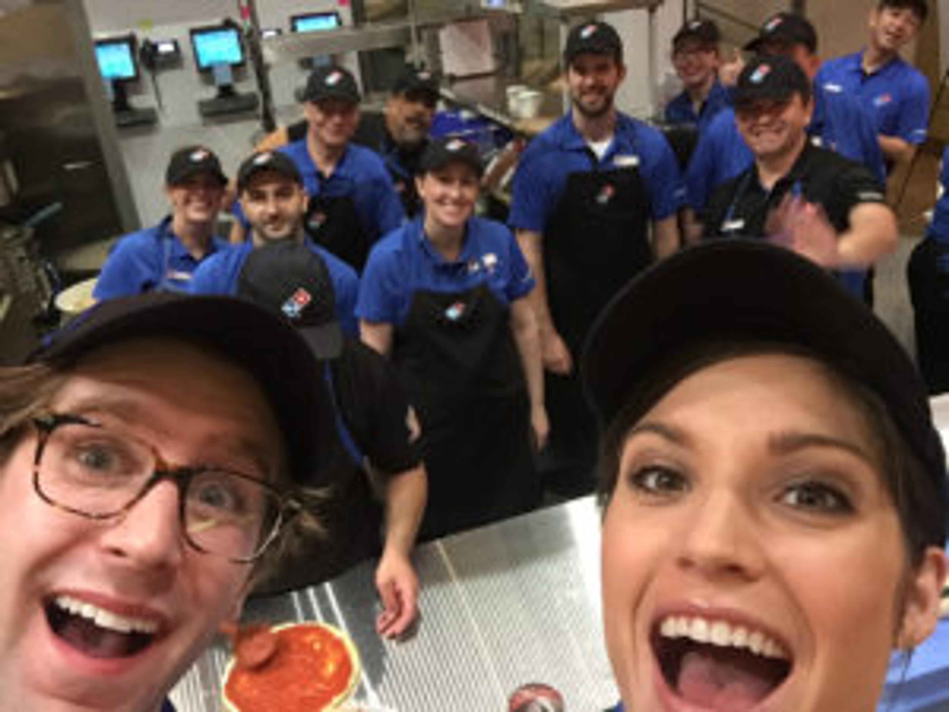 A group of Domino's employees smiles and poses for a selfie in a pizza kitchen, with one person in the foreground making a funny face. The team is wearing blue shirts and black aprons.