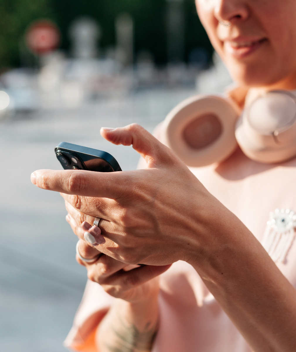 Close-up of a person outdoors using a smartphone, with wireless headphones resting around their neck.