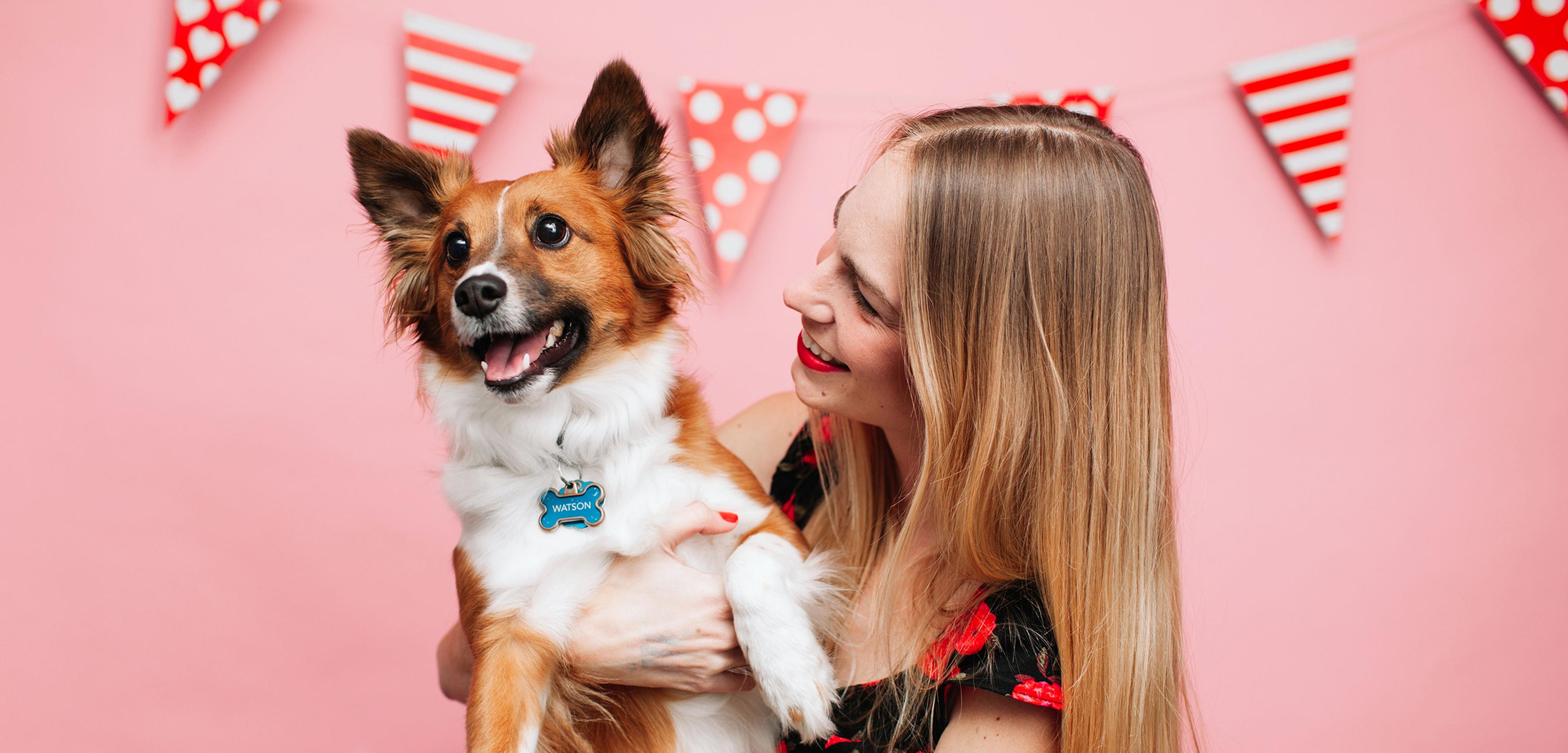 A woman with long hair smiles while holding a happy dog against a pink background decorated with red and white bunting.