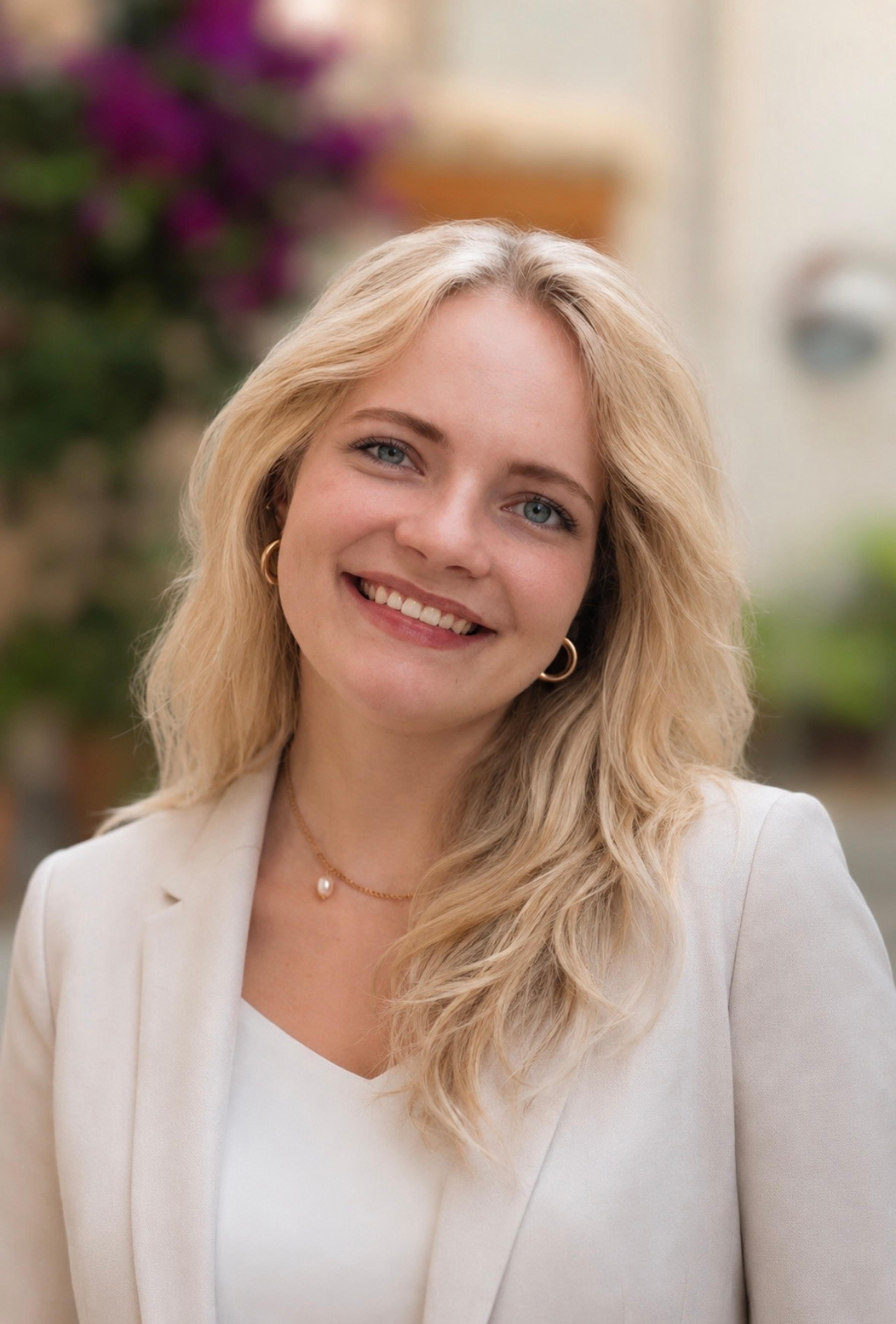 A smiling woman with long, wavy blonde hair is wearing a light-colored blazer and a white top. She stands outdoors with blurred greenery and flowers in the background.