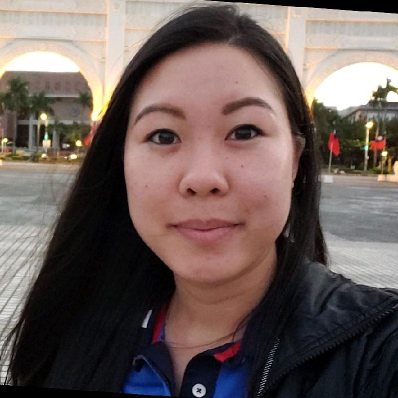 A woman with long black hair smiles at the camera, standing outdoors in a well-lit area with arches and palm trees in the background. She is wearing a dark jacket and a collared shirt.