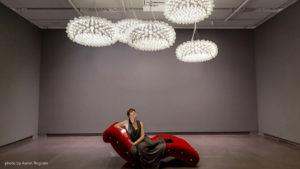 A woman sits on a red, sculptural lounge chair under modern, artistic light fixtures in a gallery space. The walls are gray, creating a minimalist backdrop.