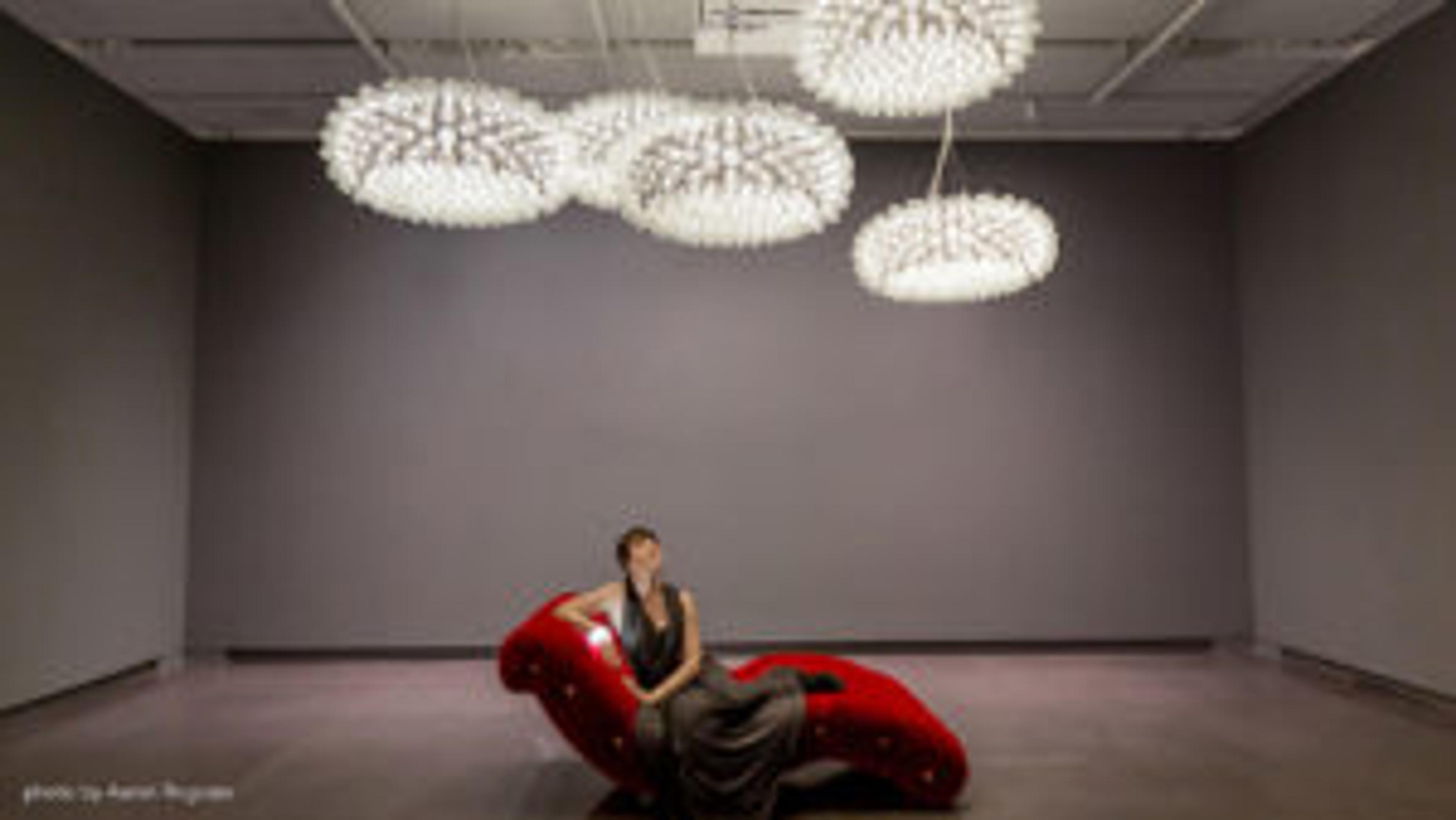 A woman sits on a red, sculptural lounge chair under modern, artistic light fixtures in a gallery space. The walls are gray, creating a minimalist backdrop.