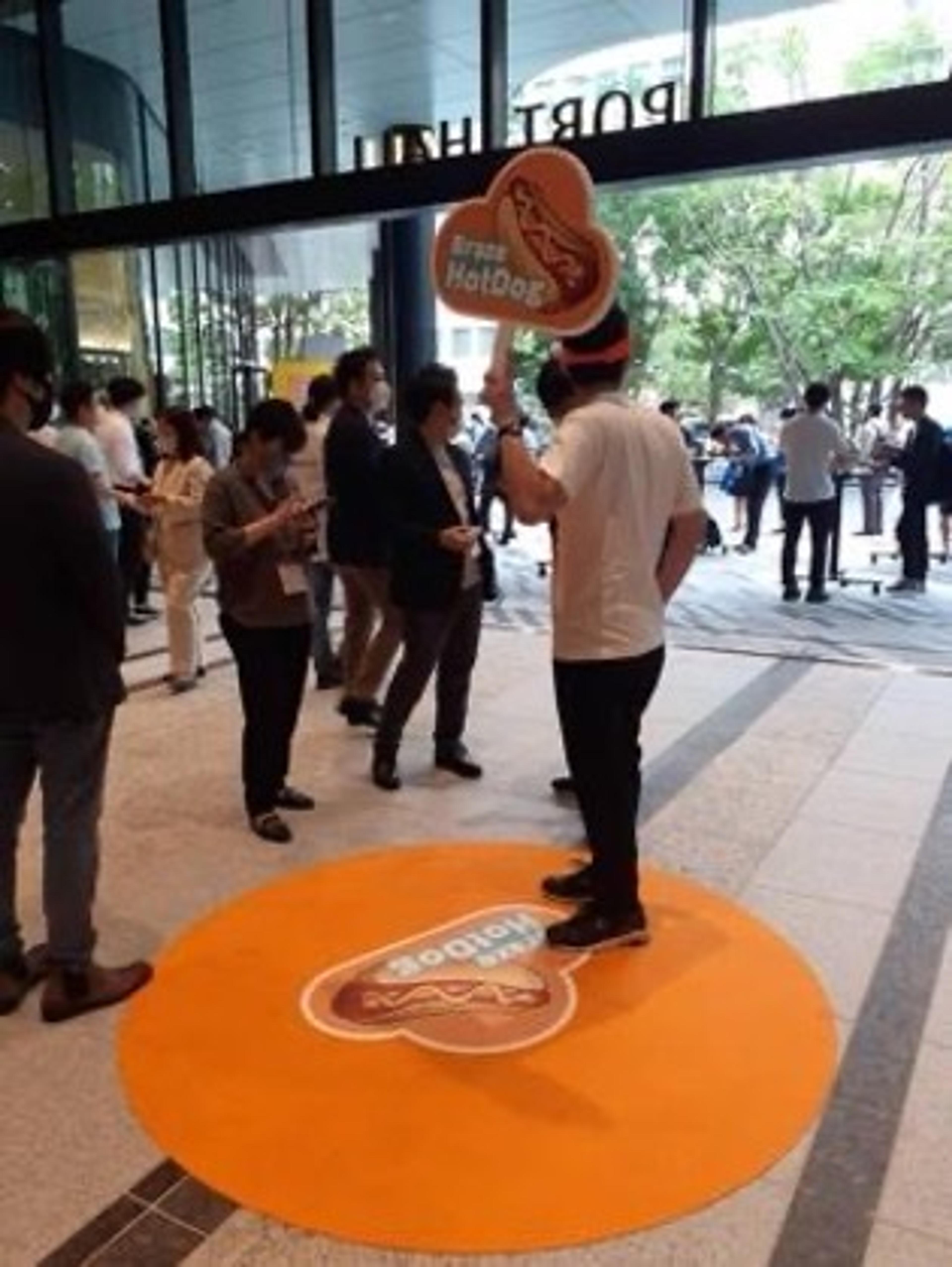 ホットドッグの看板を持った人が、イベント会場の入口付近で立っている。周囲には多くの人々が集まっている。