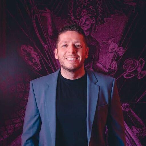 Hombre sonriente con chaqueta azul y camiseta negra, frente a un fondo oscuro con ilustraciones artísticas.