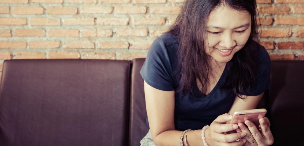 A woman sitting on a couch smiles while looking at her smartphone. The background features a brick wall.