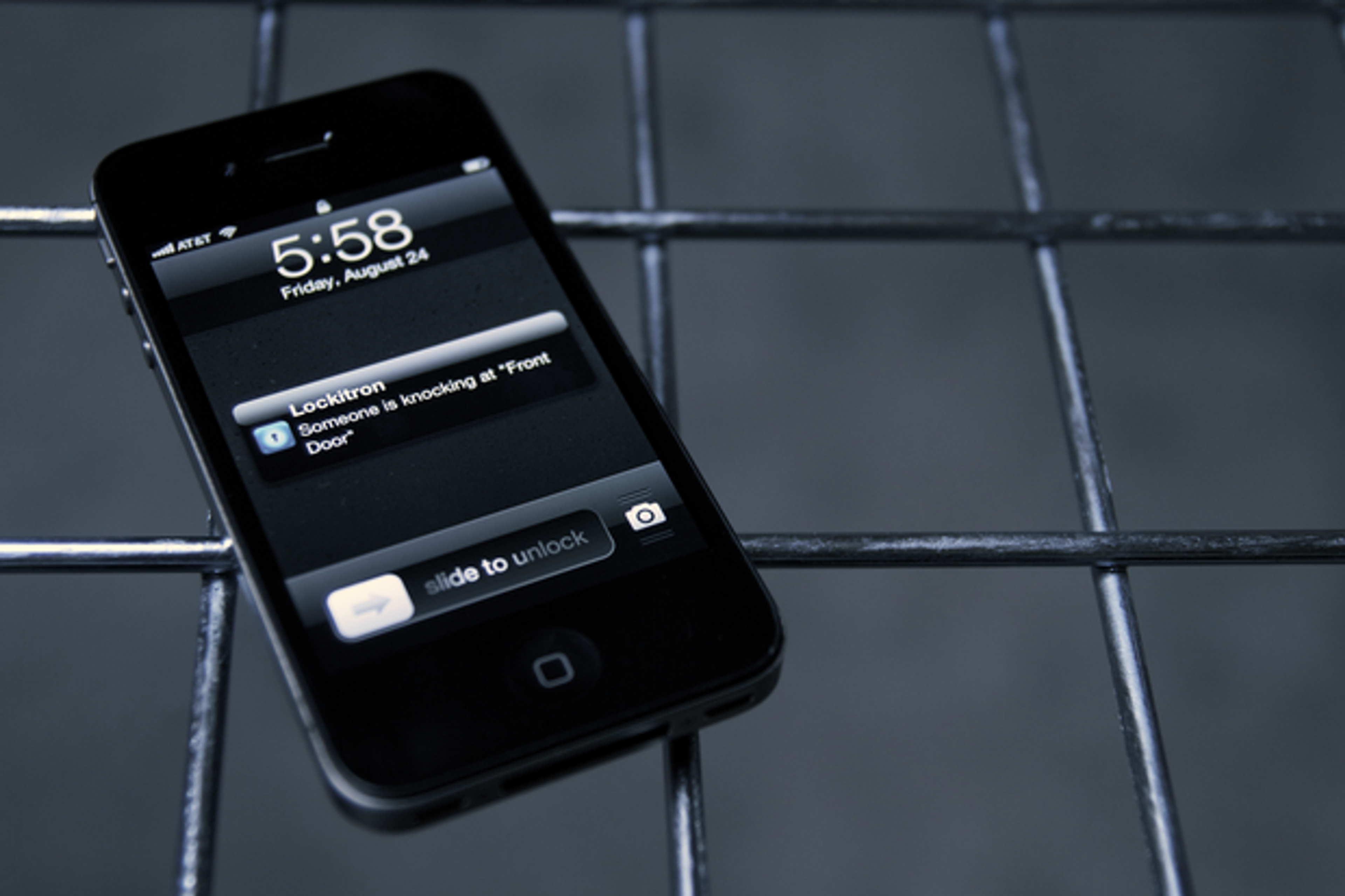 A smartphone displaying a notification that someone is knocking at the front door, with the time showing 5:58 PM on August 24. The phone is resting on a metal grid surface.
