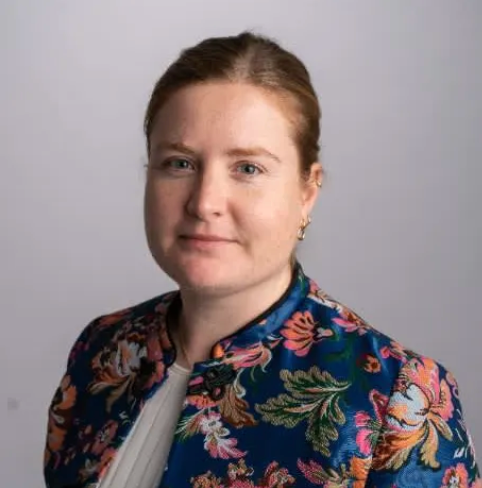 A woman with auburn hair wearing a floral-patterned jacket poses for a portrait against a neutral background. She has a slight smile and is looking directly at the camera.