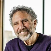 A smiling man with curly gray hair and a beard, wearing a purple sweater, is posed in front of a window with a blurred background.