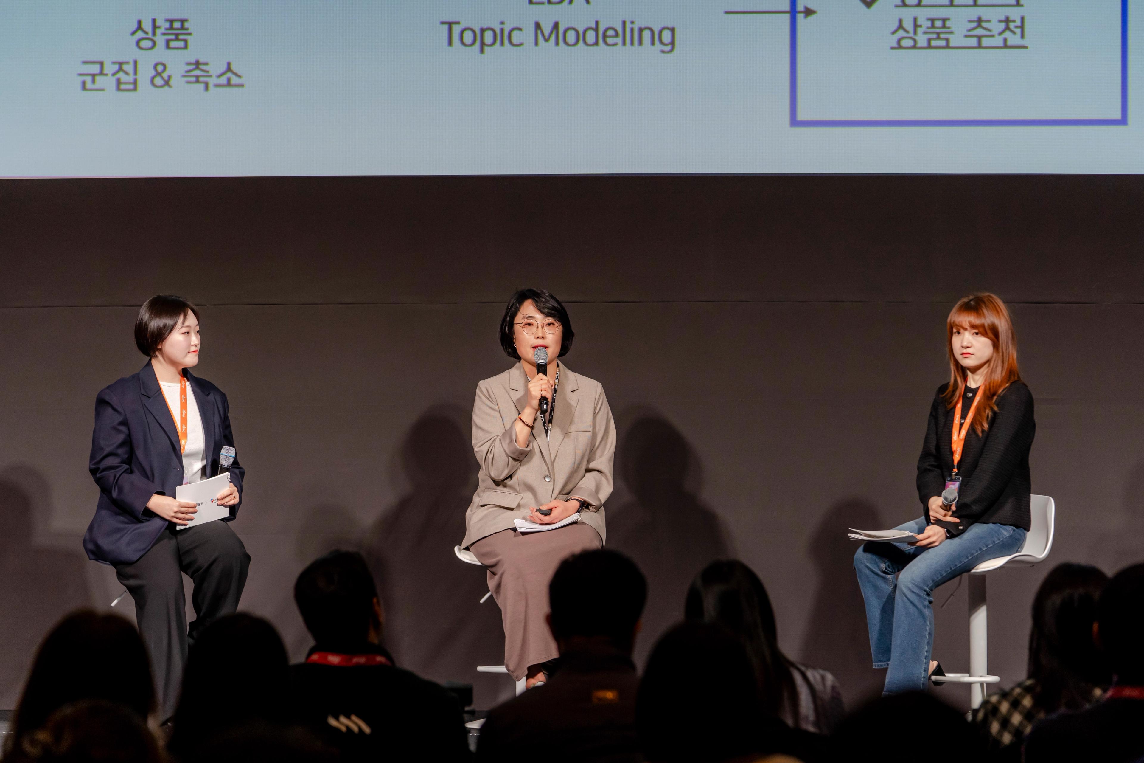 three women are sitting on a stage talking to a crowd .