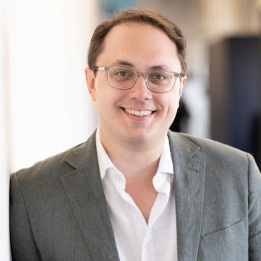 A smiling man with glasses is leaning against a wall, wearing a gray blazer over a white shirt. The background is softly blurred, suggesting an office environment.