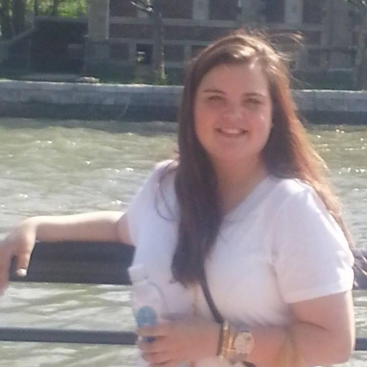 Smiling woman with long brown hair sitting on a bench by the water.