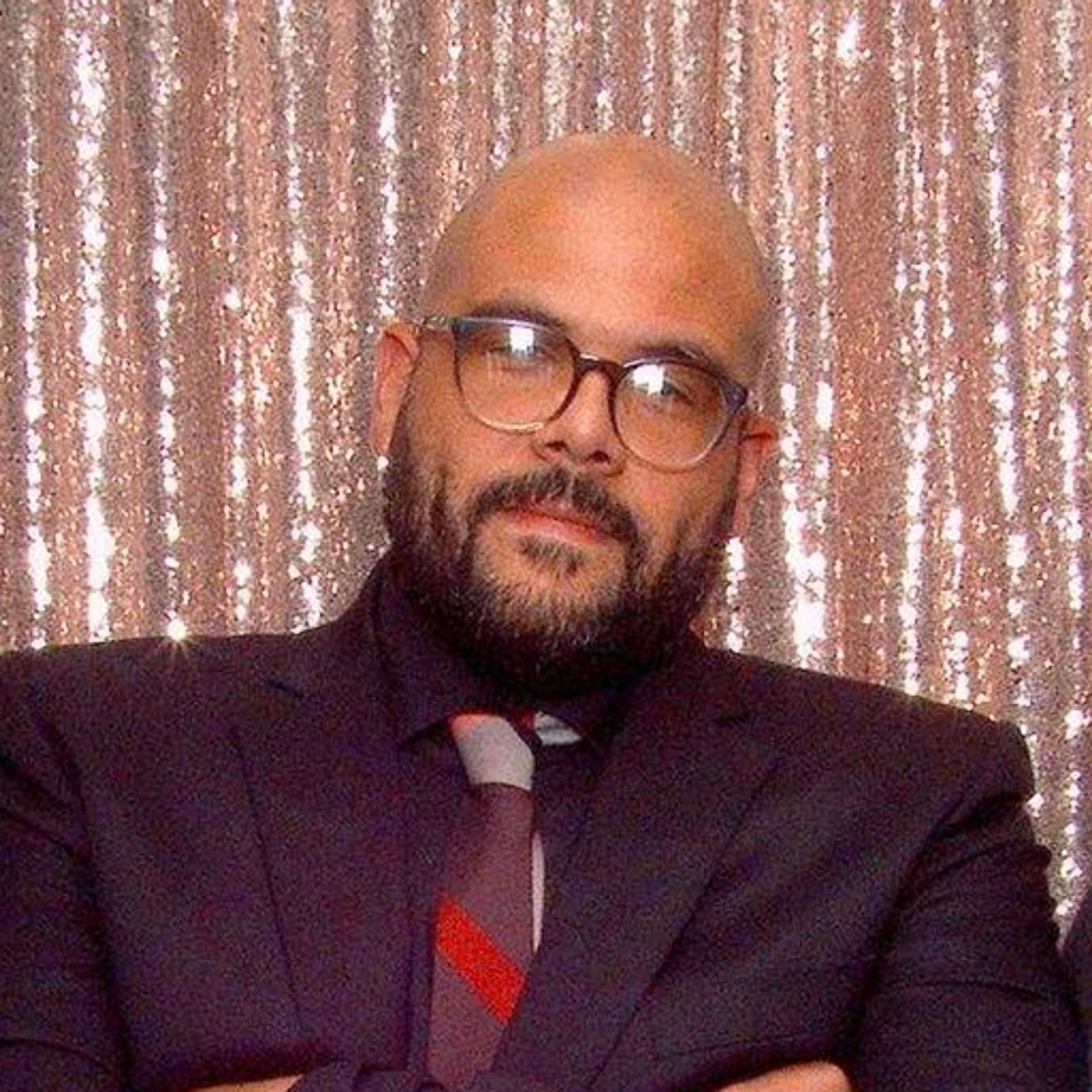 Headshot of a bald man with glasses and a beard, wearing a dark suit and patterned tie against a shimmering backdrop.z