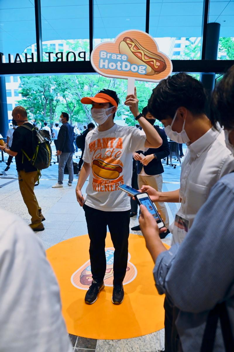ホットドッグの看板を持ったスタッフが、イベント会場で参加者にアピールしています。周囲には他の参加者がいます。