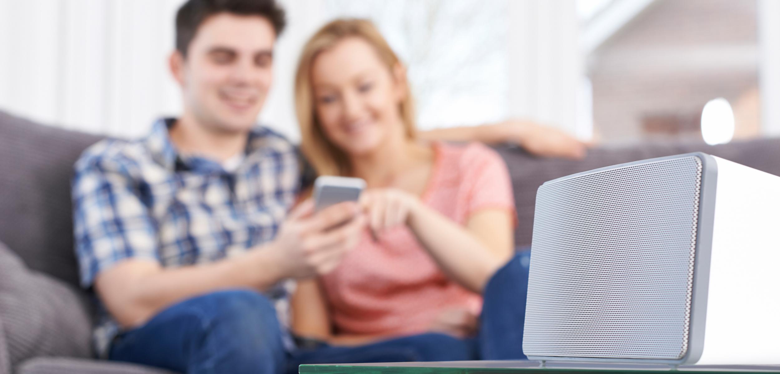 A couple sits on a couch, smiling and looking at a smartphone, with a speaker on a glass table in the foreground.