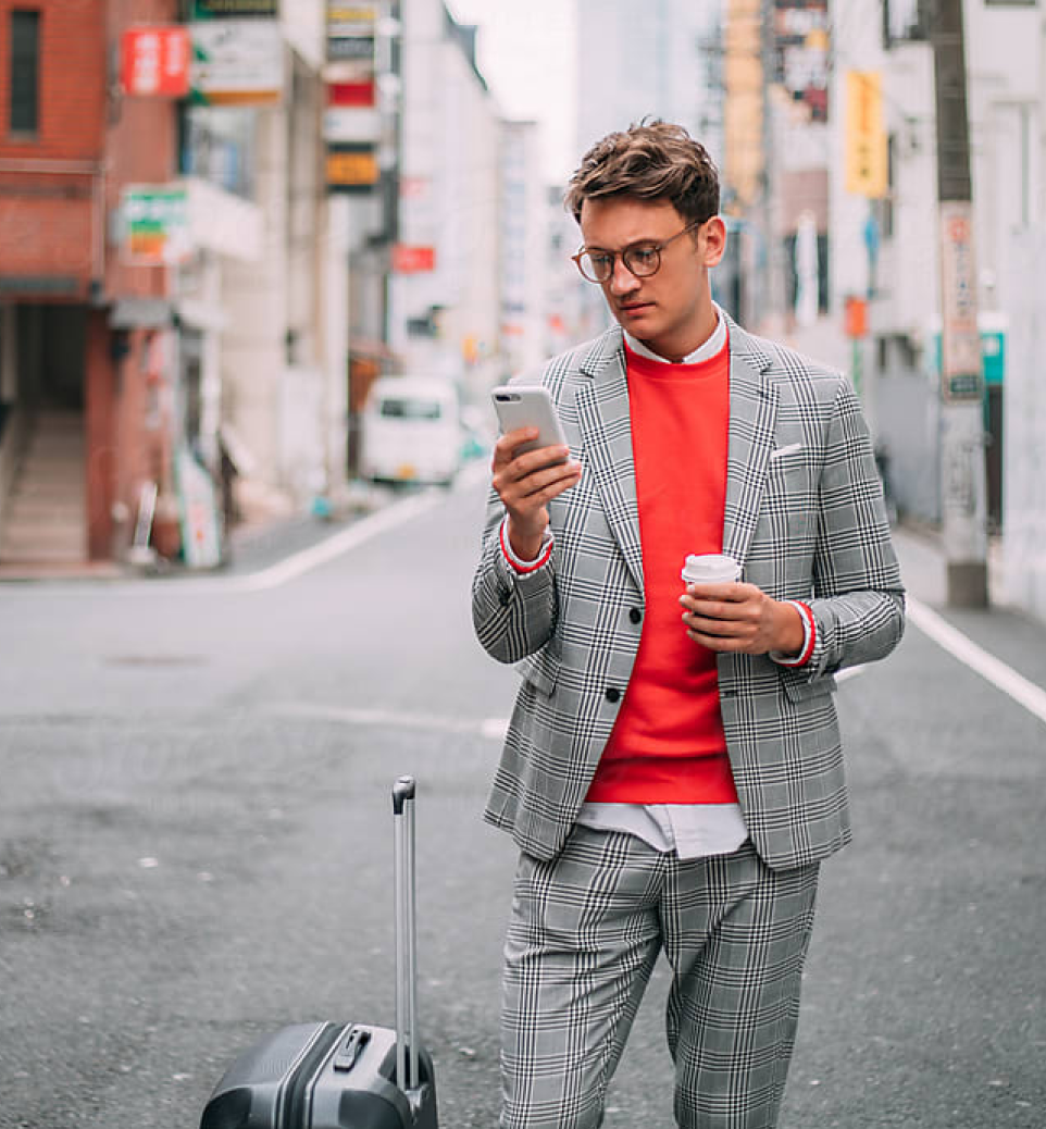 A man in a checked suit and red sweater checks his phone while holding a coffee cup, standing on a city street with a suitcase beside him.