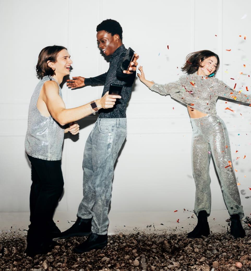 Three stylishly dressed friends in sparkly outfits laugh and dance together as colorful confetti falls around them.