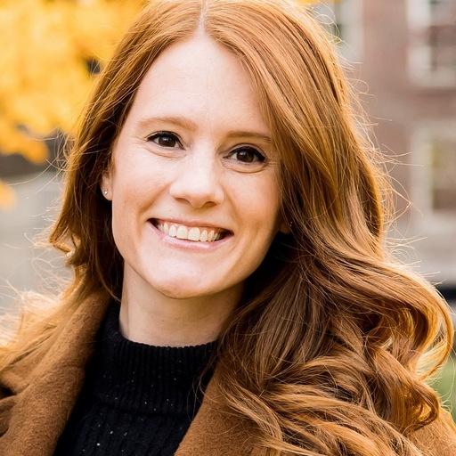 A smiling woman with long, wavy red hair stands outdoors, surrounded by autumn leaves. She is wearing a dark sweater and a brown coat.