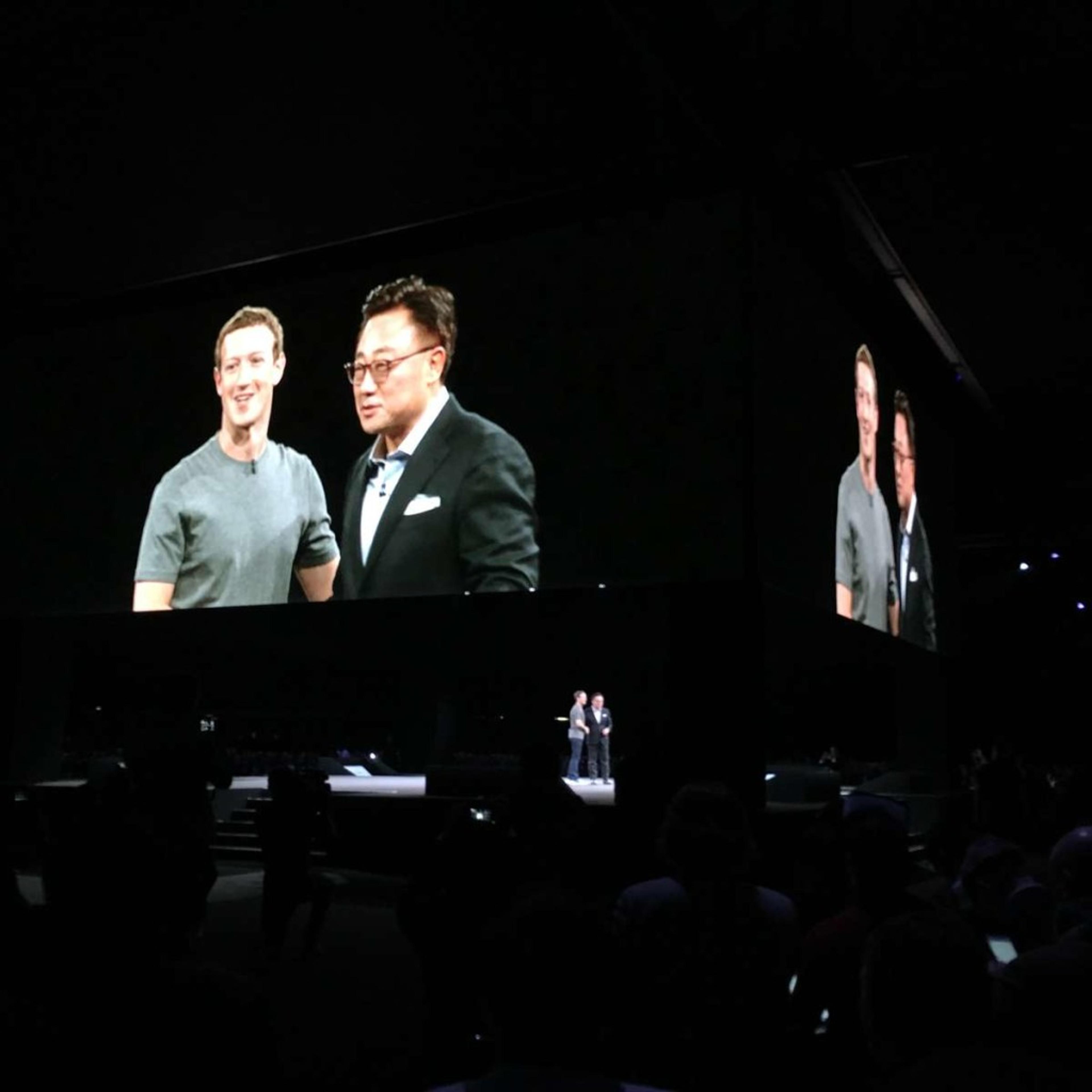 A large screen displays two men on stage at an event, with one man wearing a gray shirt and the other in a suit. The audience is visible in the foreground.