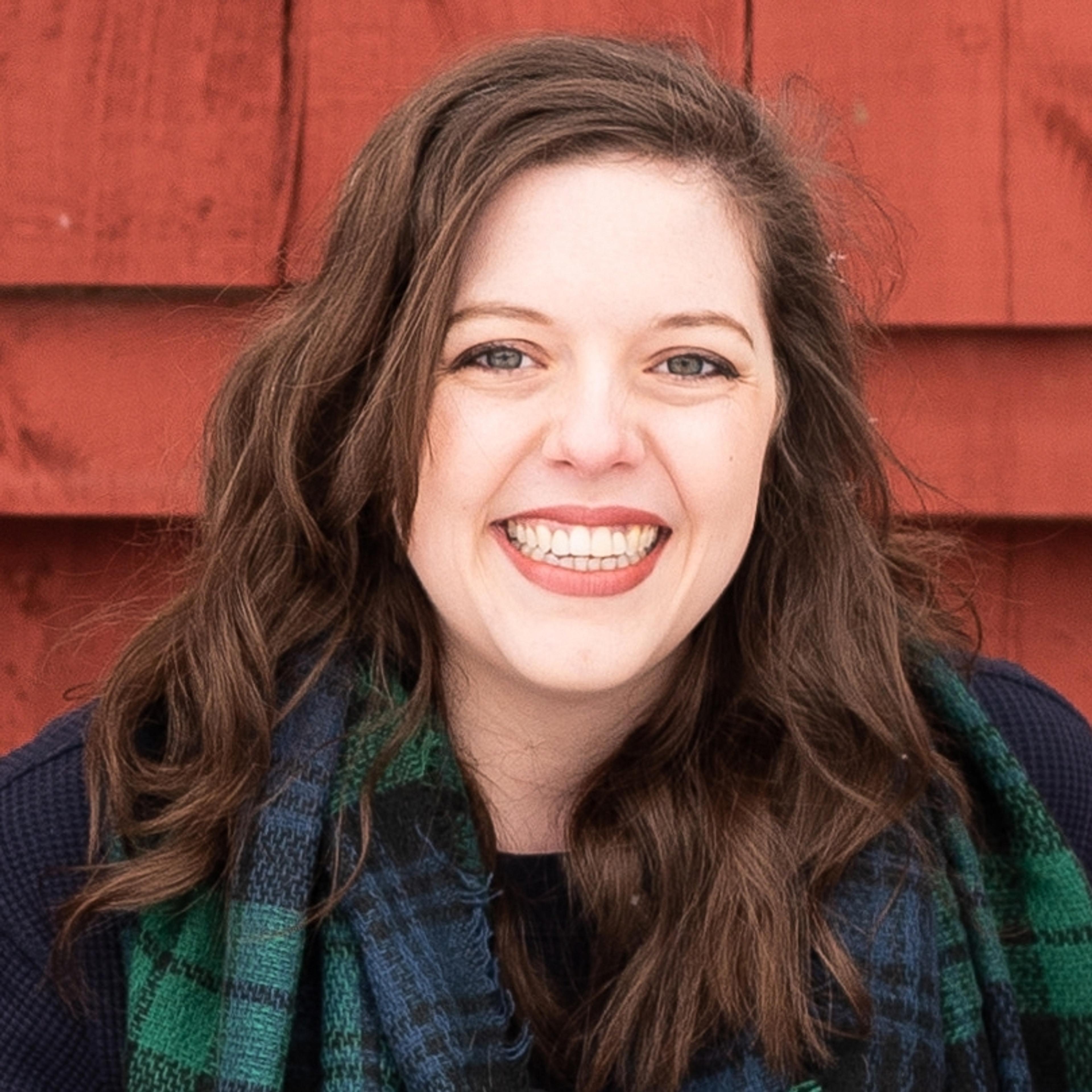A smiling woman with long, wavy brown hair is wearing a blue sweater and a green plaid scarf. She is in front of a red wooden background.