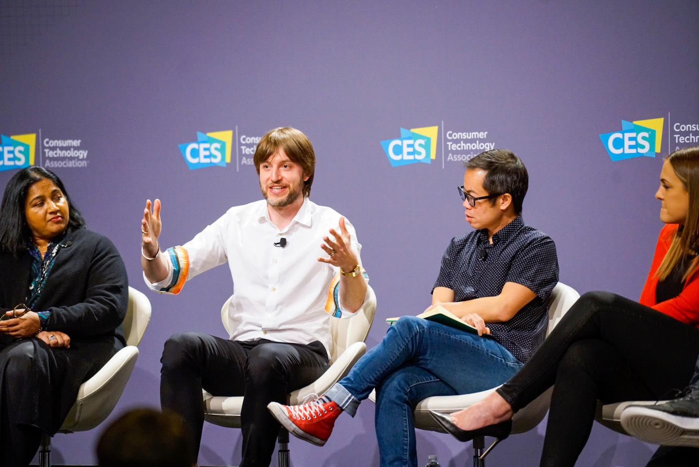 A panel discussion at CES featuring four speakers. One speaker, wearing a white shirt and red shoes, is animatedly gesturing while speaking.