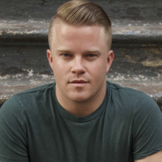 A young man with short, styled hair is sitting on a set of stairs, wearing a green t-shirt and looking directly at the camera. The background features a textured wall and worn steps.
