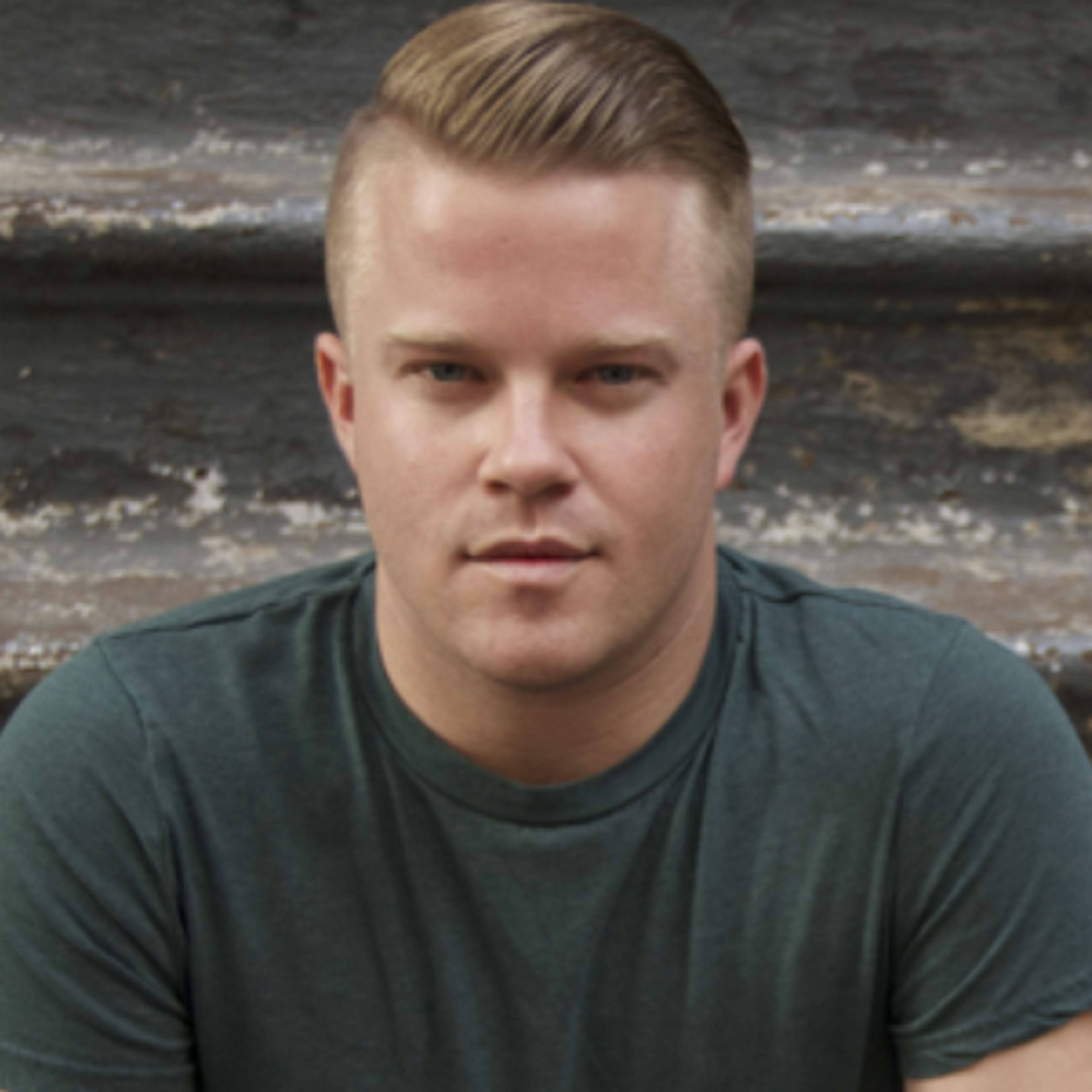 A young man with short, styled hair is sitting on a set of stairs, wearing a green t-shirt and looking directly at the camera. The background features a textured wall and worn steps.
