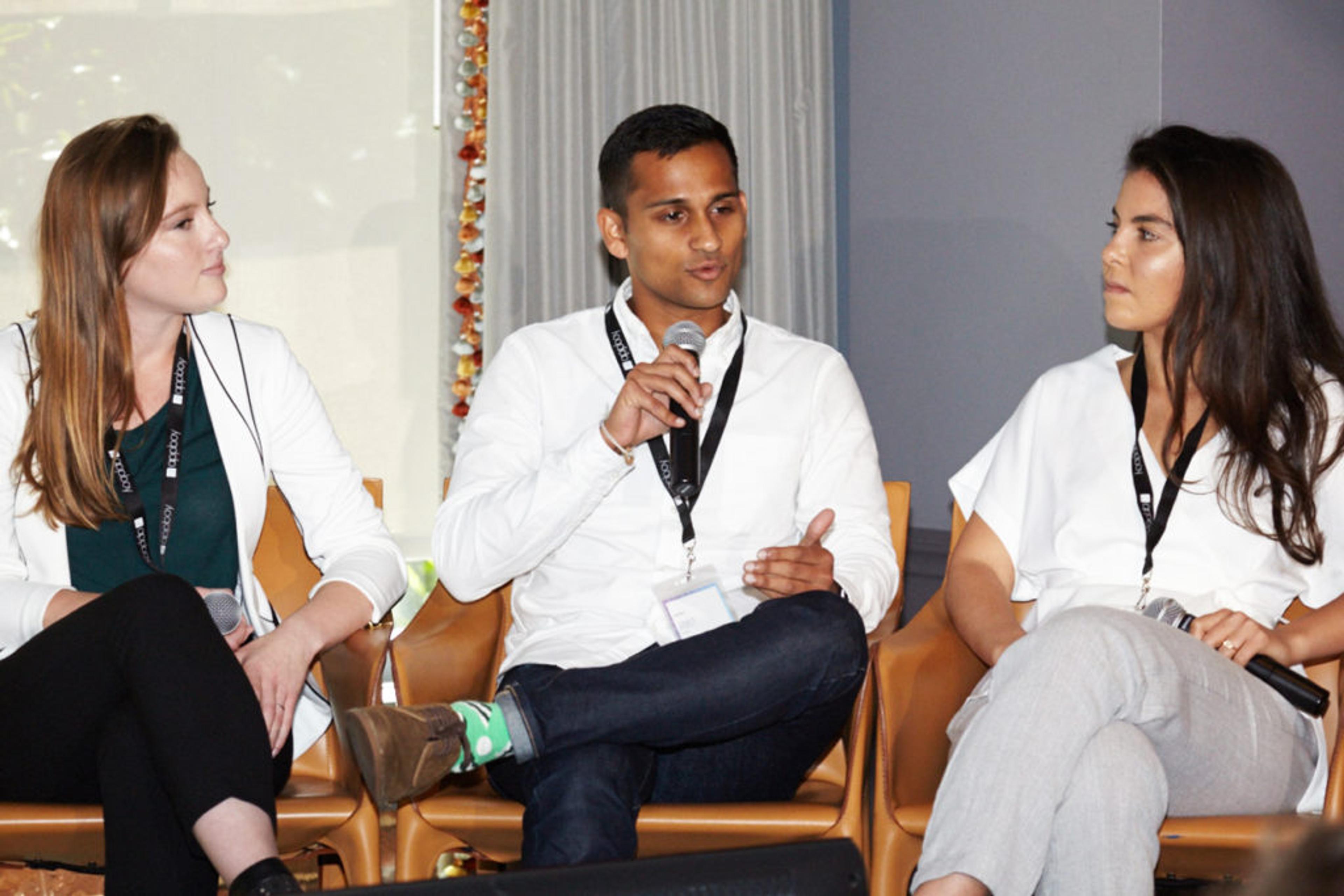 Three individuals sit on a panel discussion, with one person speaking into a microphone while the others listen attentively. They are dressed in casual business attire.
