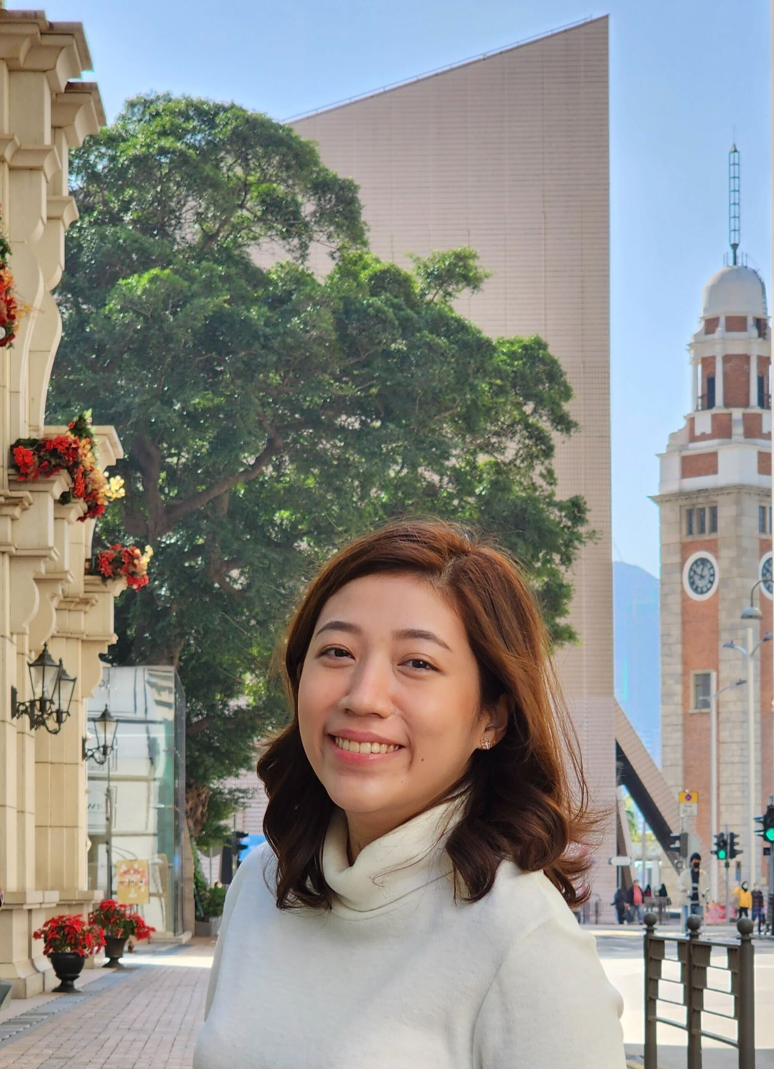 A woman with shoulder-length brown hair smiles in front of a large tree and modern buildings. Flowers decorate the area, adding color to the scene.