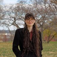 Persona con cabello largo y oscuro, usando gafas y un abrigo negro, de pie en un parque con árboles al fondo.