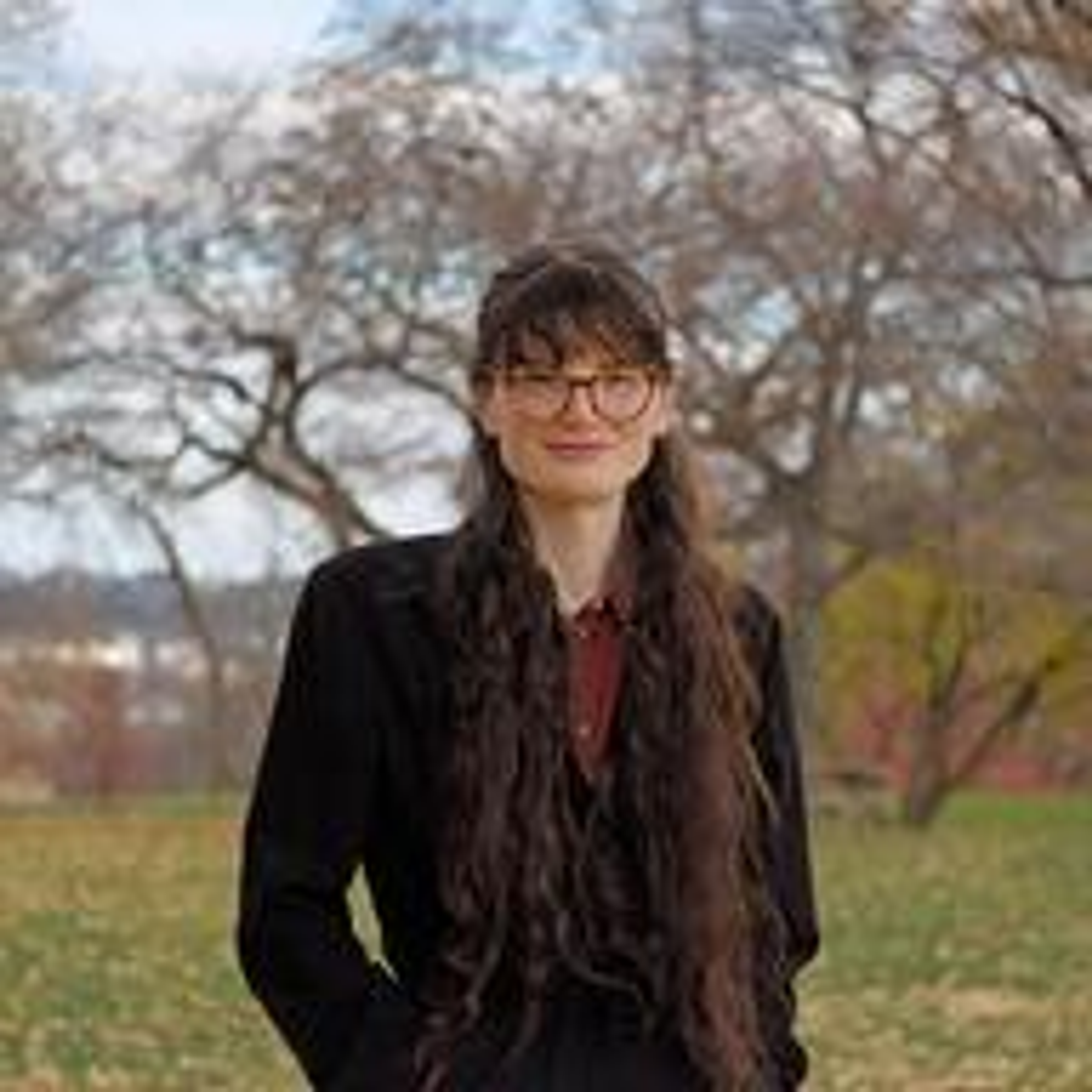 Persona con cabello largo y oscuro, usando gafas y un abrigo negro, de pie en un parque con árboles al fondo.